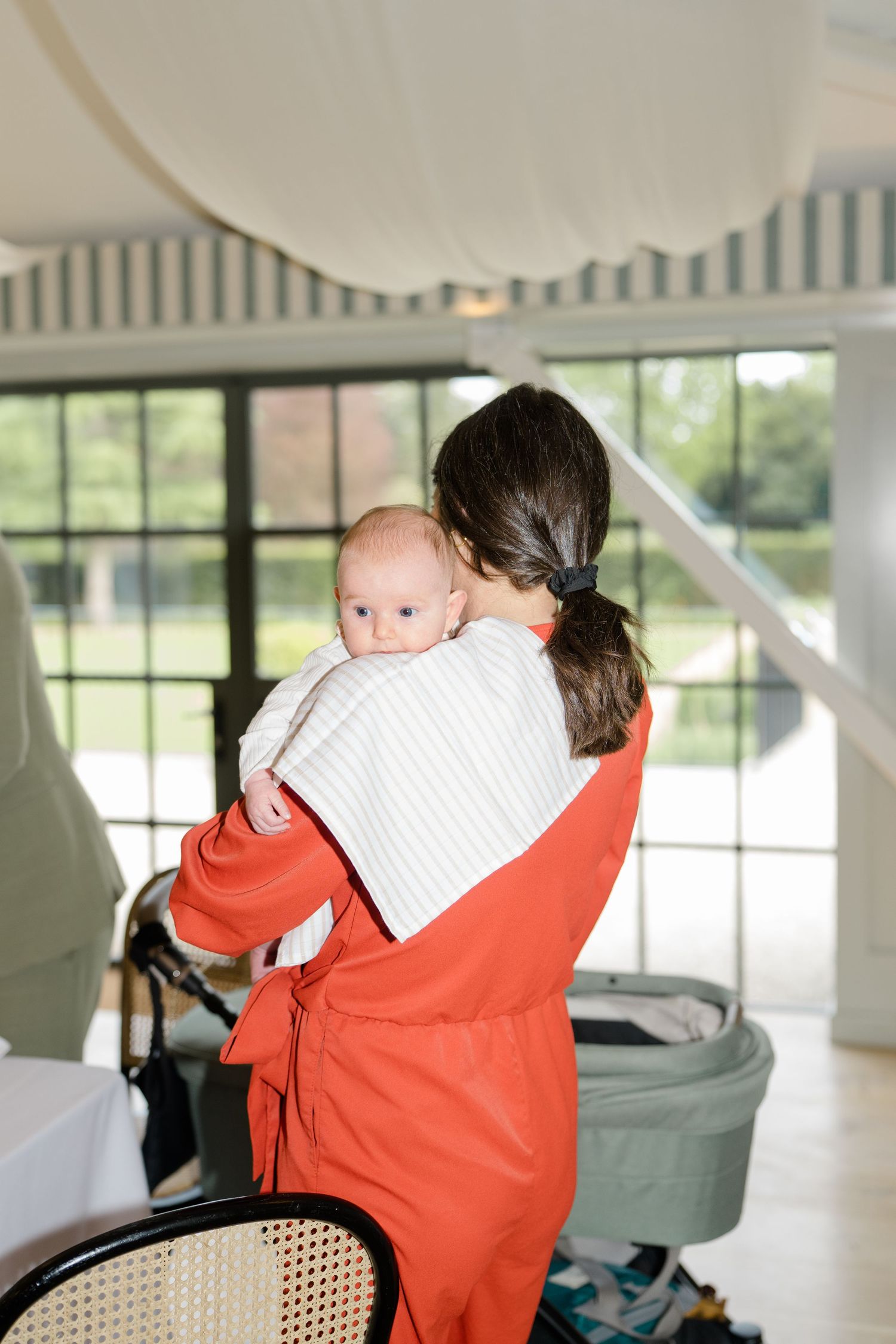 A wedding guest in red dress holds and comforts a newborn baby near a pram in a bright marquee venue.
