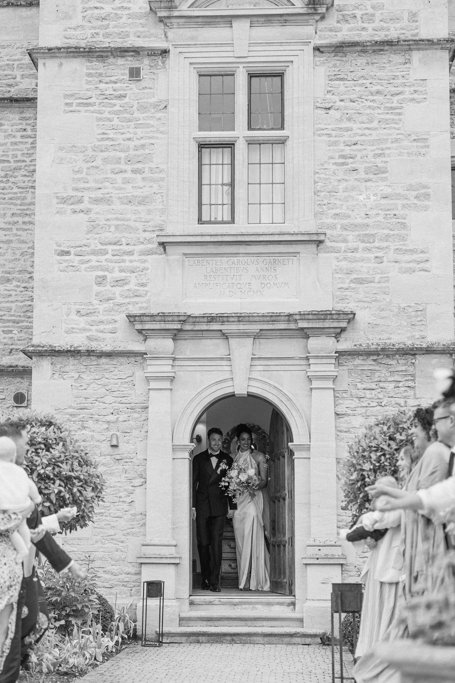 Newlyweds emerging from an arched doorway of a historic stone building greeted by cheering wedding guests.