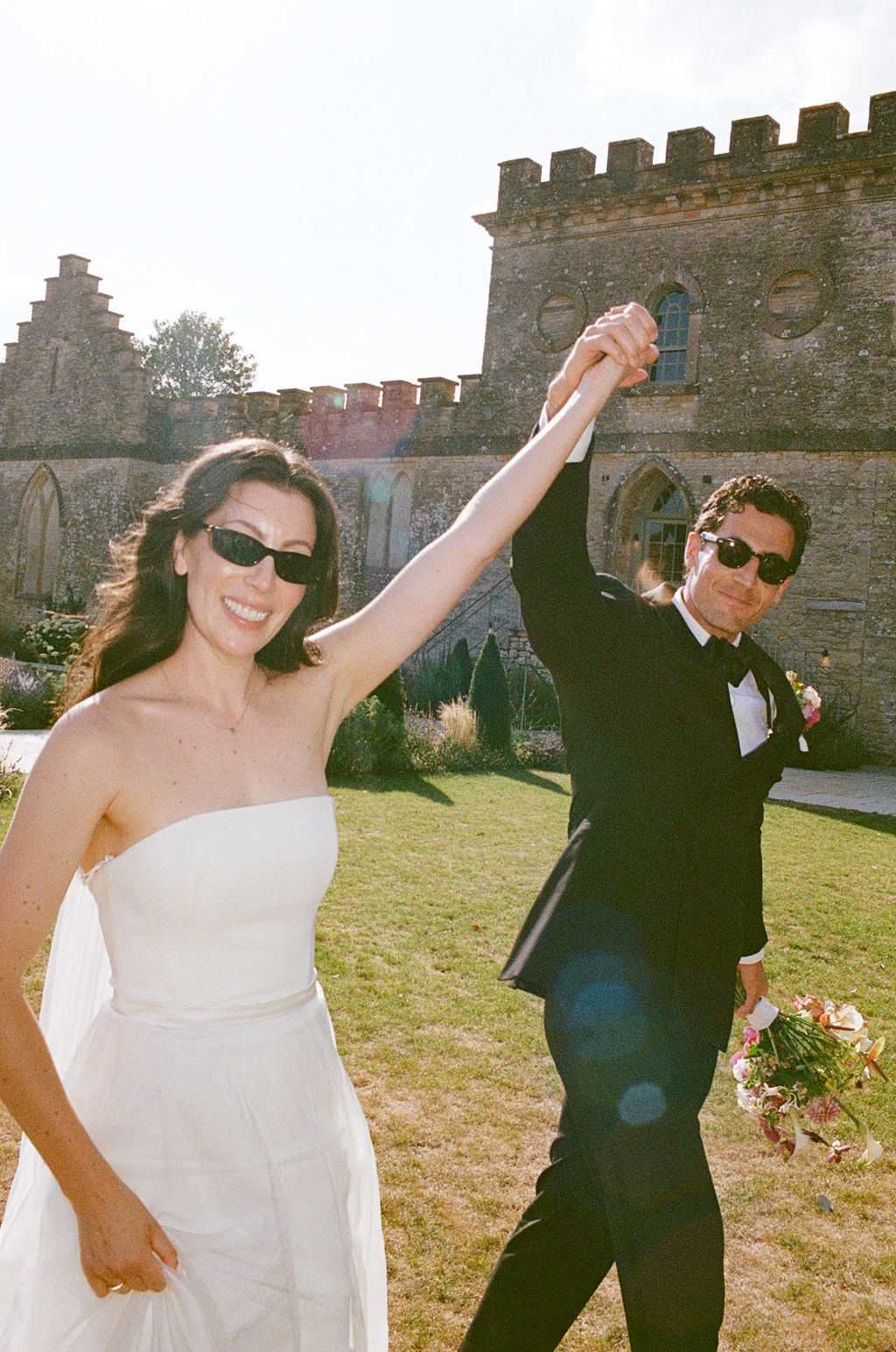 Joyful bride and groom in sunglasses raising hands together outside a historic stone castle on their wedding day.