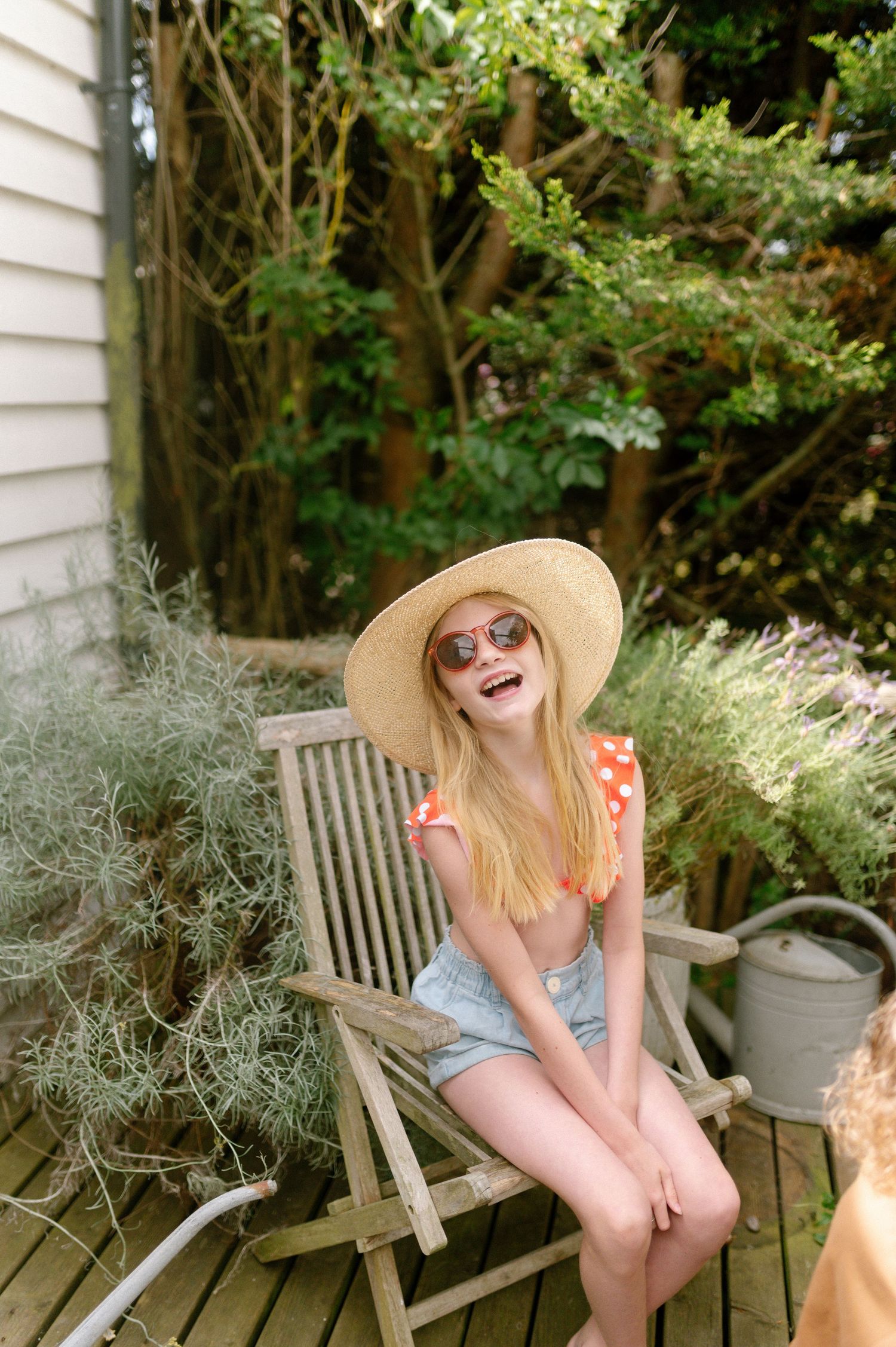 A person in a sun hat sits laughing on a wooden garden chair surrounded by lush greenery.