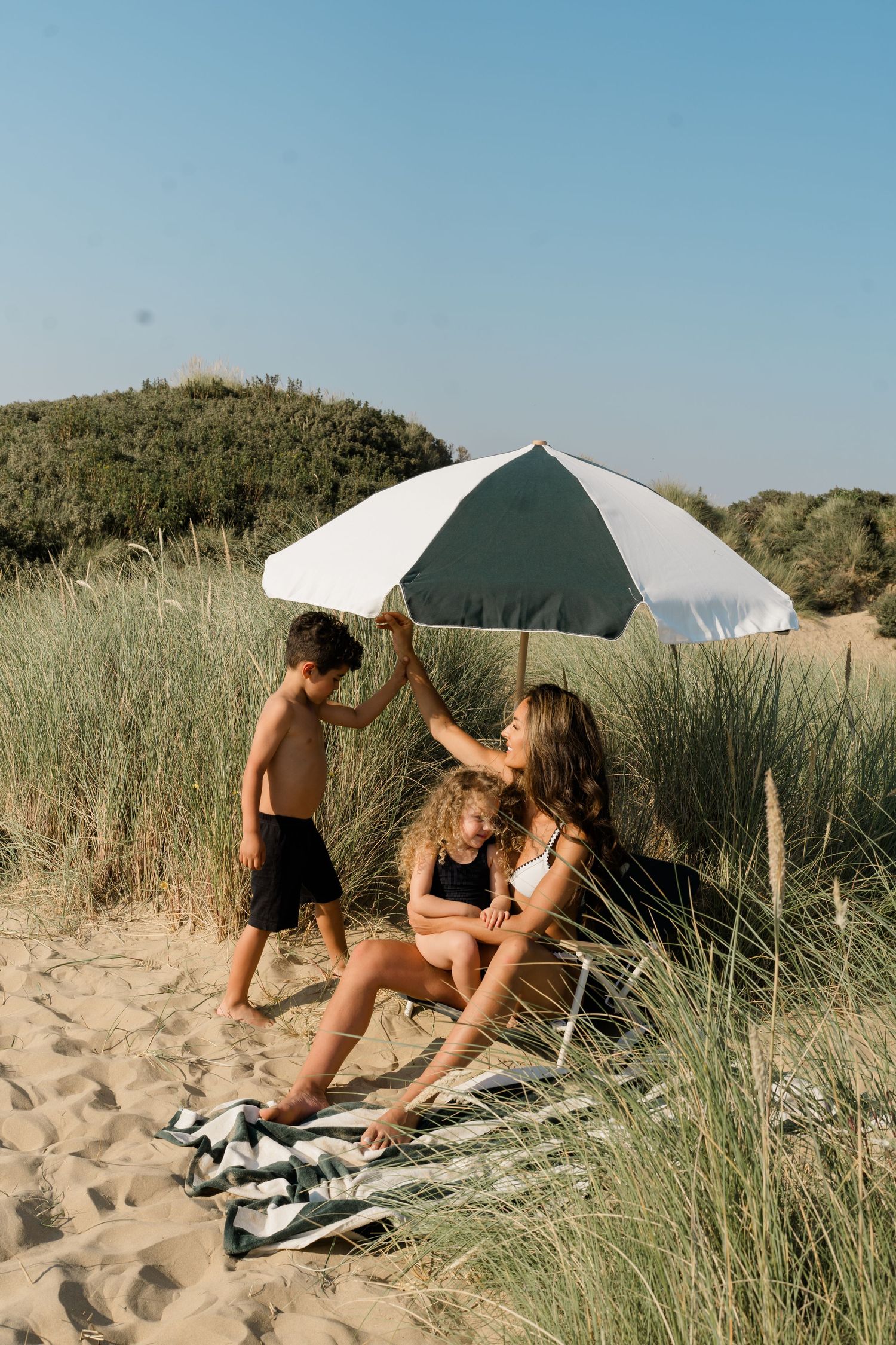 Friends lounging on striped towel under beach umbrella among coastal dunes.