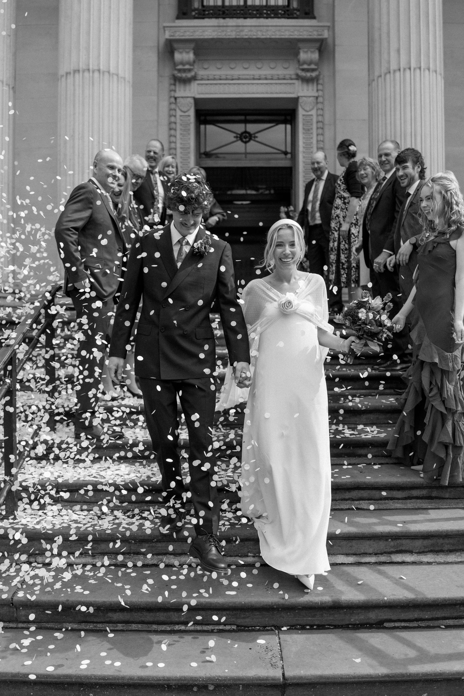 Black and white photo of wedding celebration with confetti against classical architecture.
