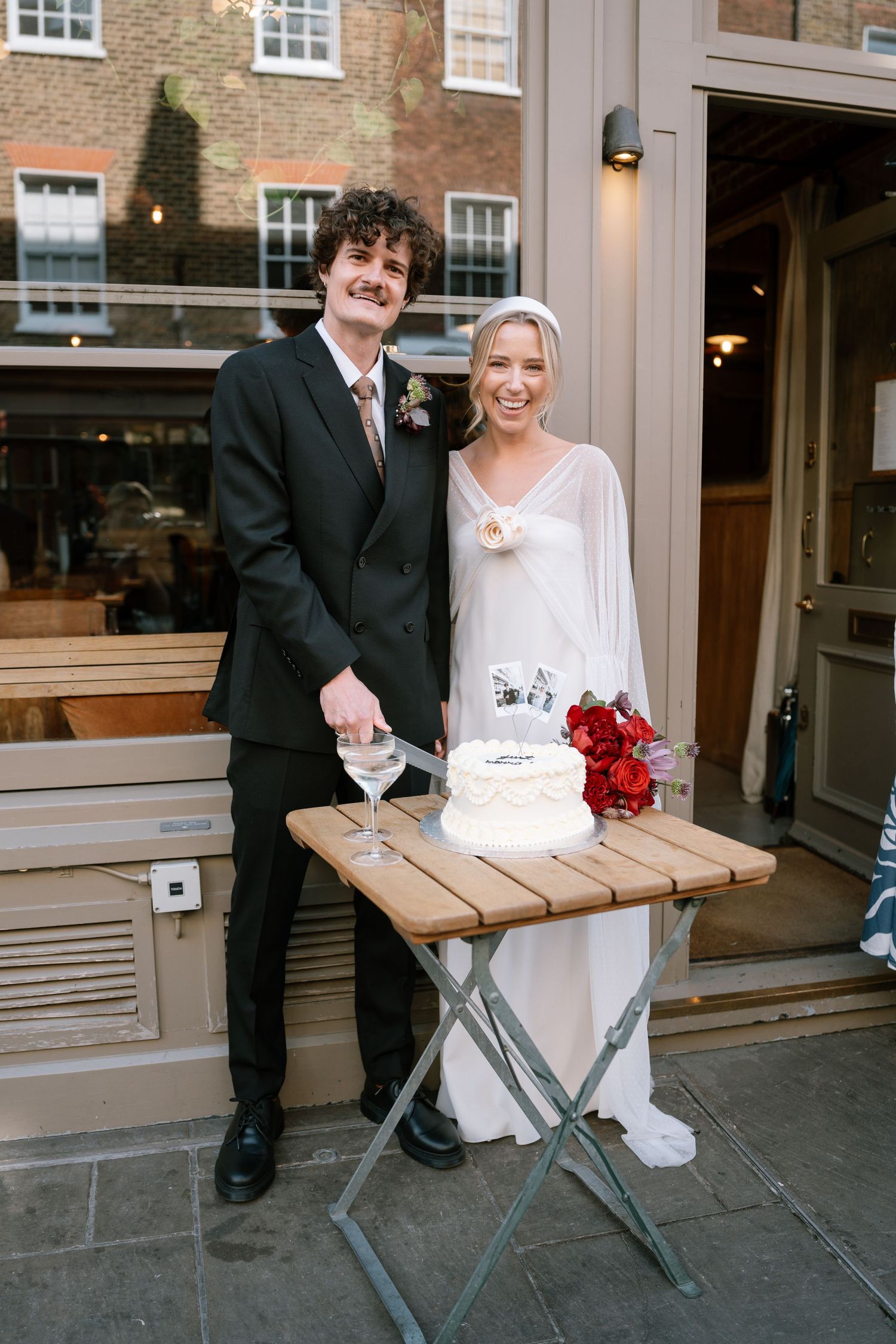 A newlywed couple poses with their wedding cake and red roses on an outdoor patio.