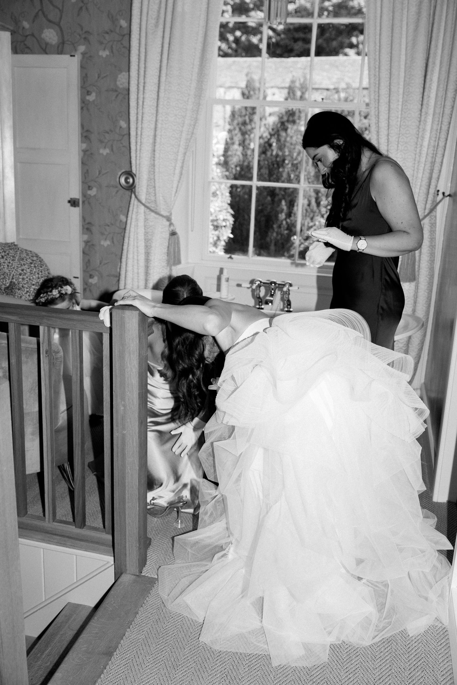 A black and white photo of wedding preparations showing a bride in a flowing dress getting assistance.