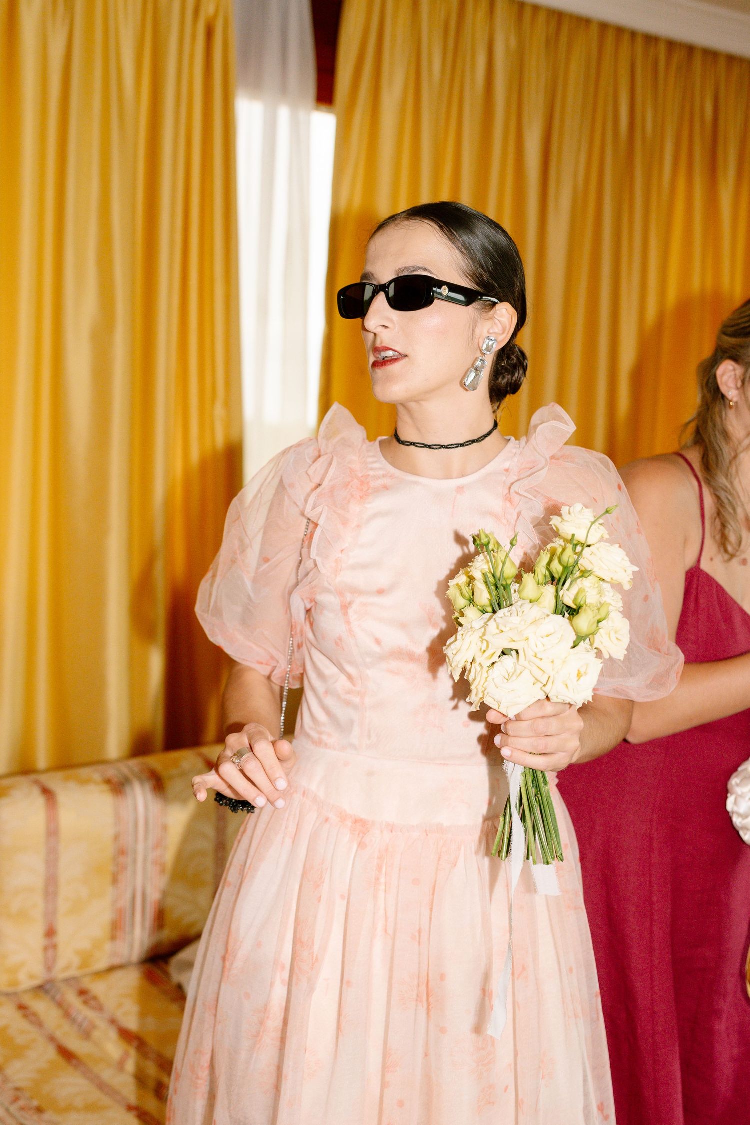 Person in a peach dress holding a bouquet against yellow curtains.