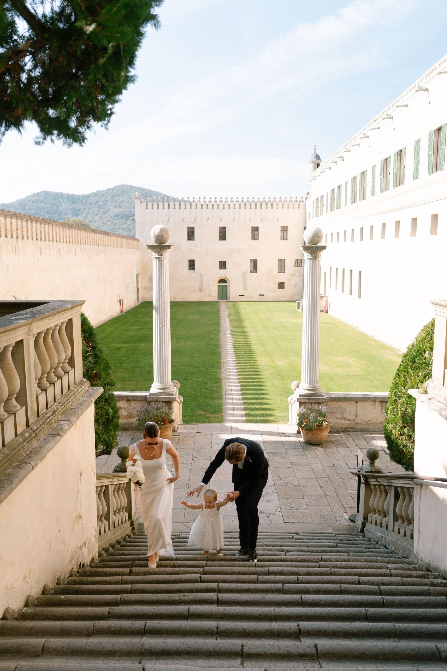 Wedding party descends grand stone staircase at historic castle with mountain views.