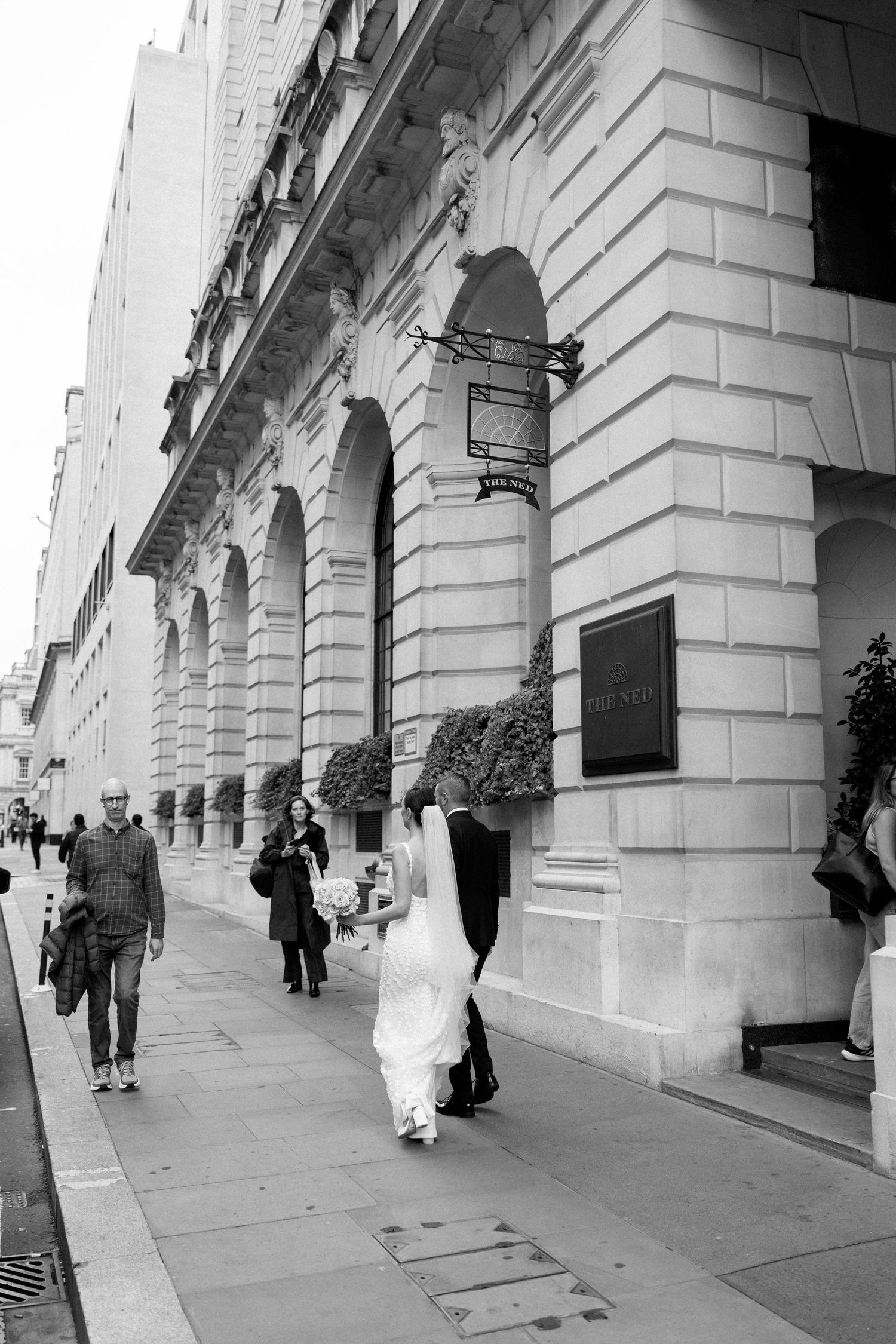 Black and white wedding photograph showing a couple walking along a classical columned building facade.