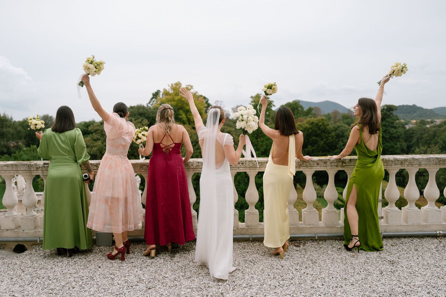 Wedding party in colorful dresses faces away showing bouquets against mountain backdrop.
