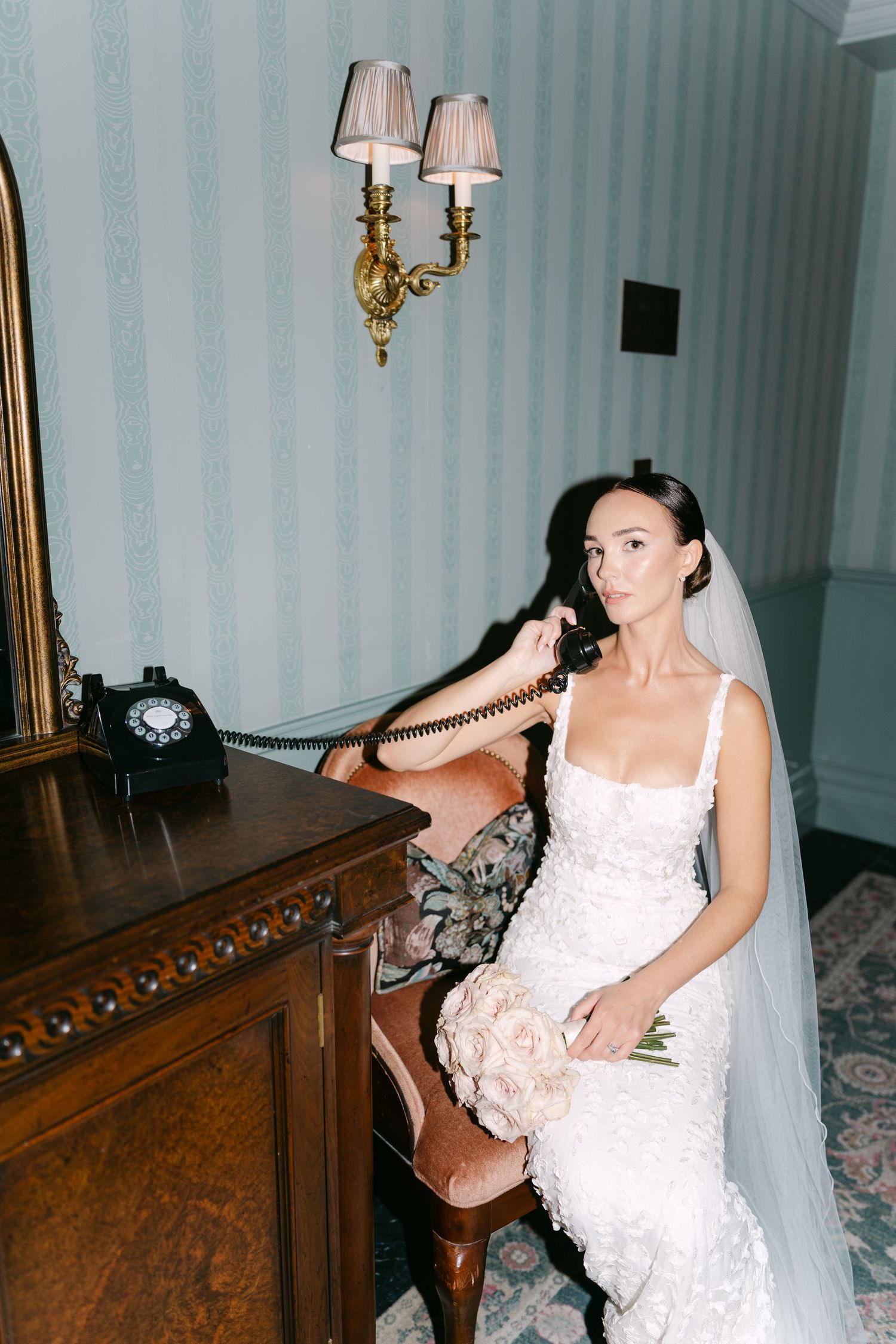 Bride in white lace gown holds bouquet near vintage wooden desk.