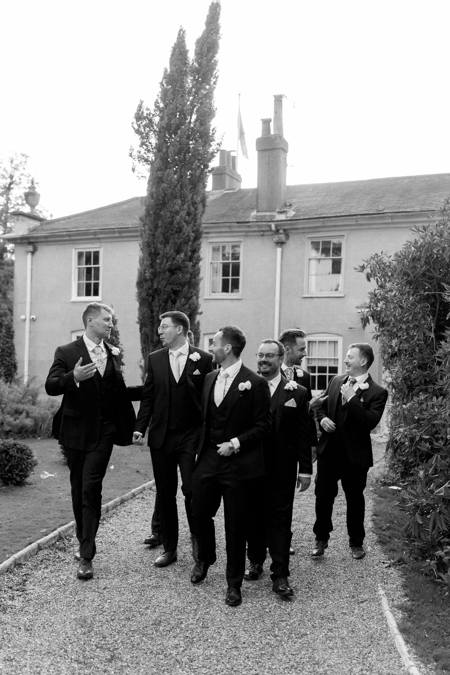 Wedding party in formal attire walking and laughing together on a garden path in black and white.