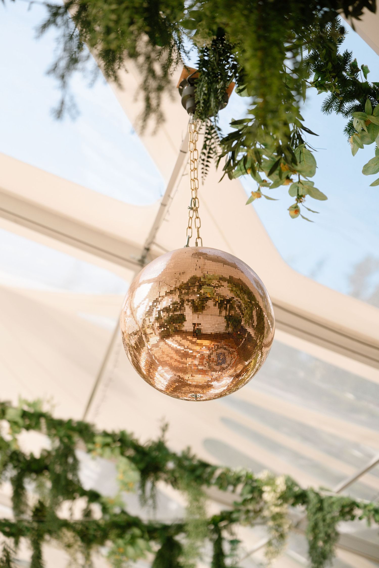 Rose gold spherical decorative lights hanging from clear tent ceiling with greenery.