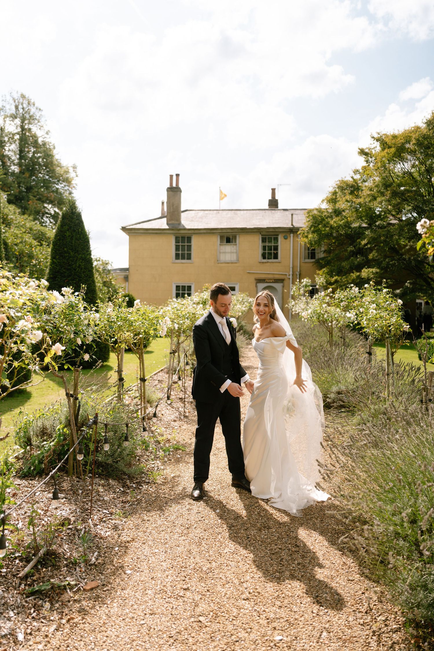 Newlyweds walk together on a gravel path surrounded by blooming flowers and lavender.