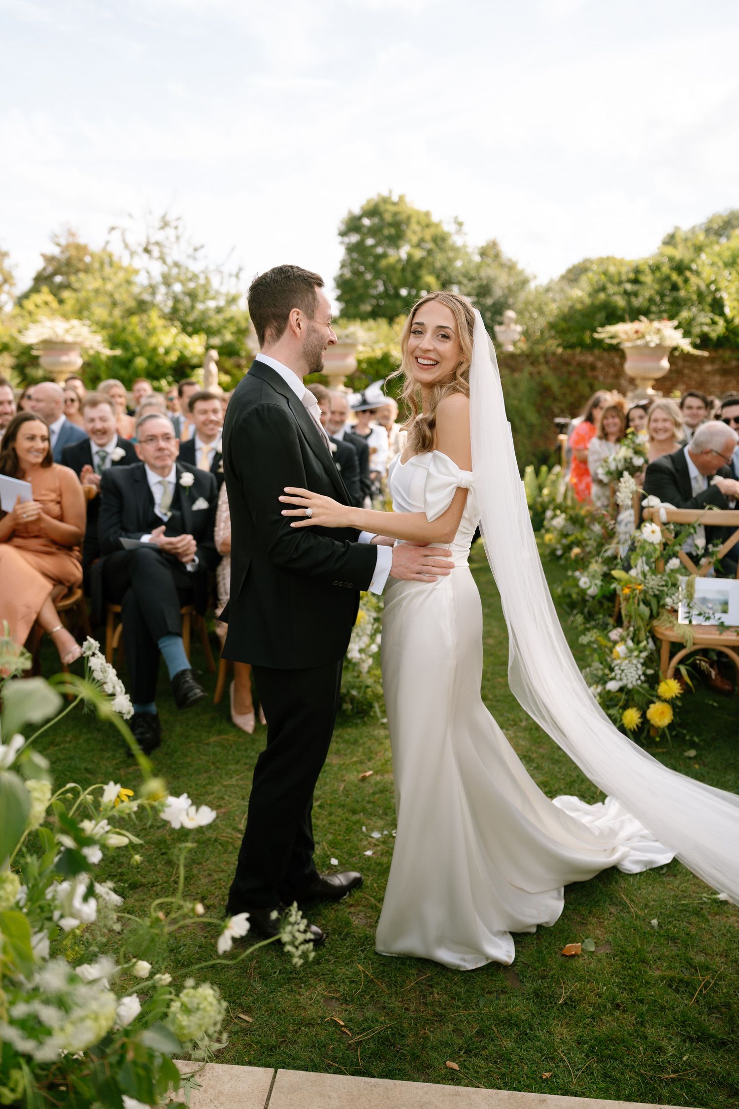 Bride in flowing white gown and groom share a moment during outdoor garden ceremony.