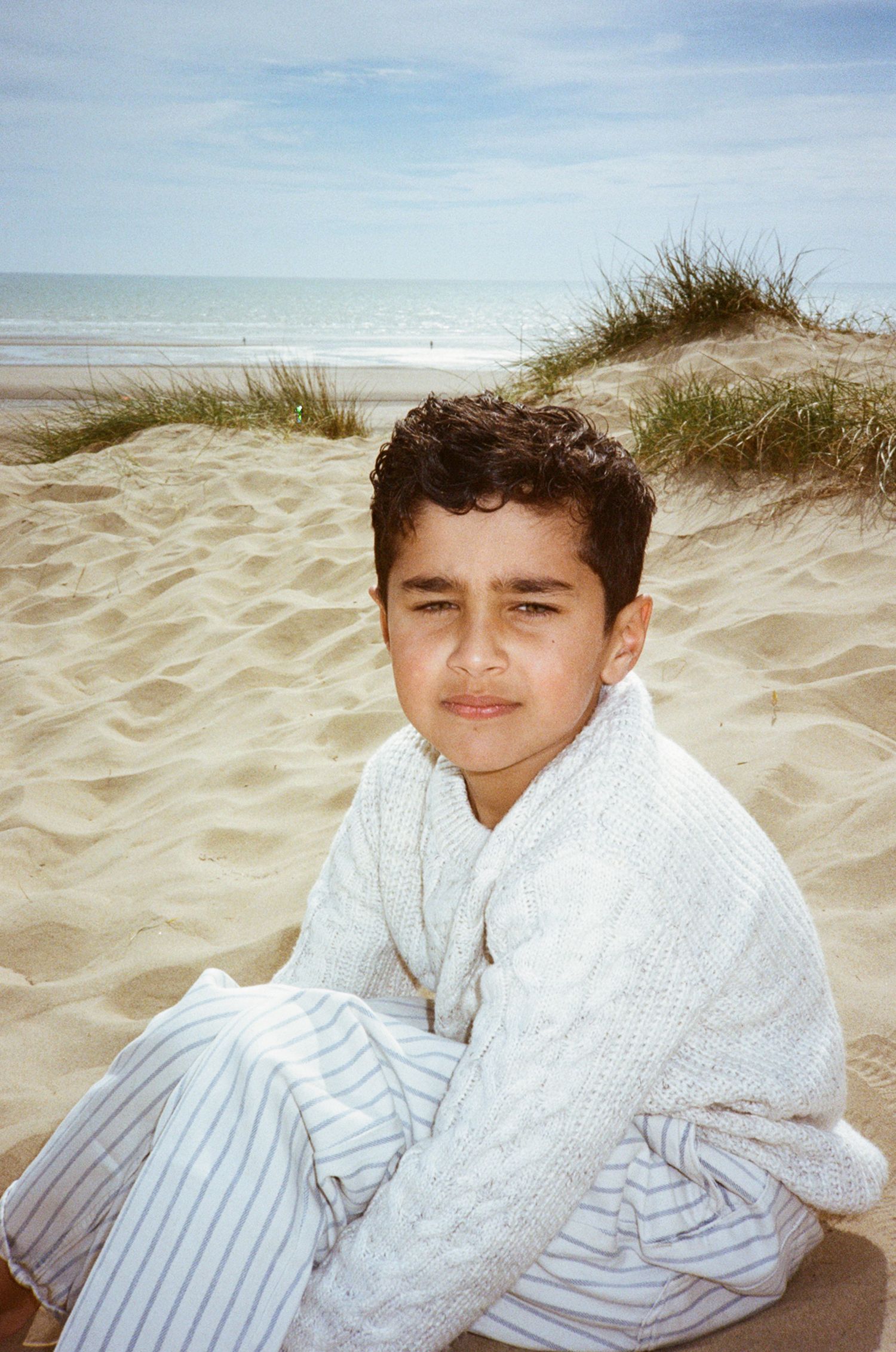 A young boy in white knitwear sits on sandy beach dunes with ocean waves and blue sky in the background.