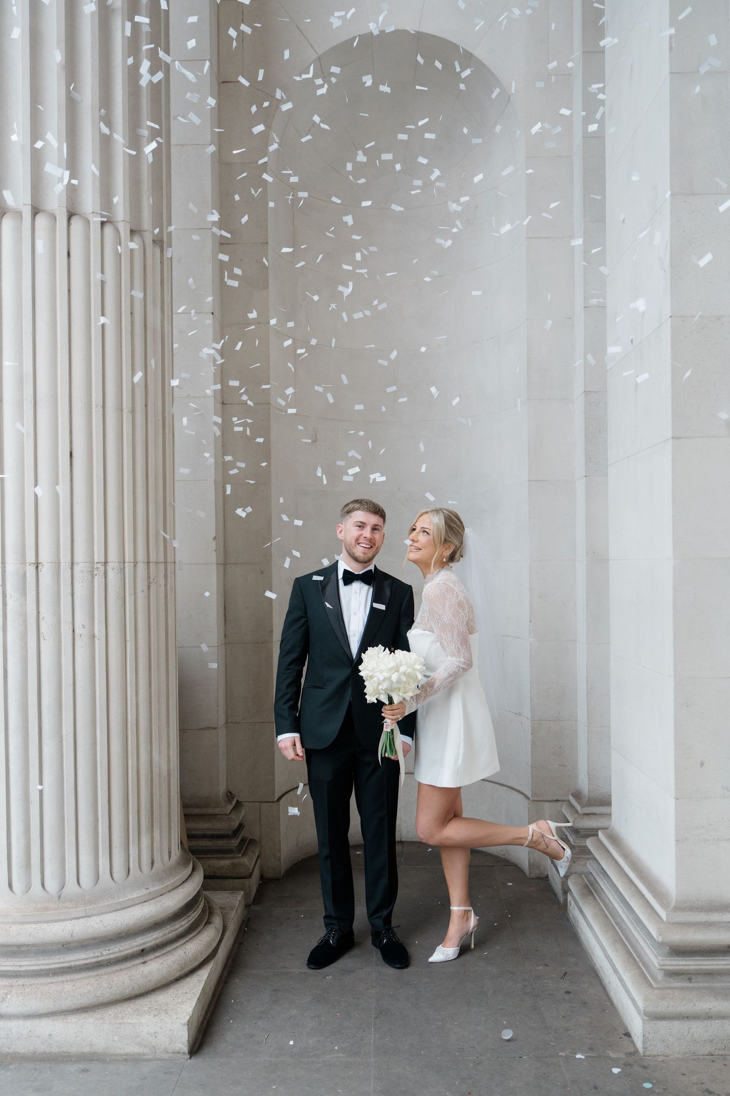 A wedding couple poses between classical white columns as confetti falls around them.