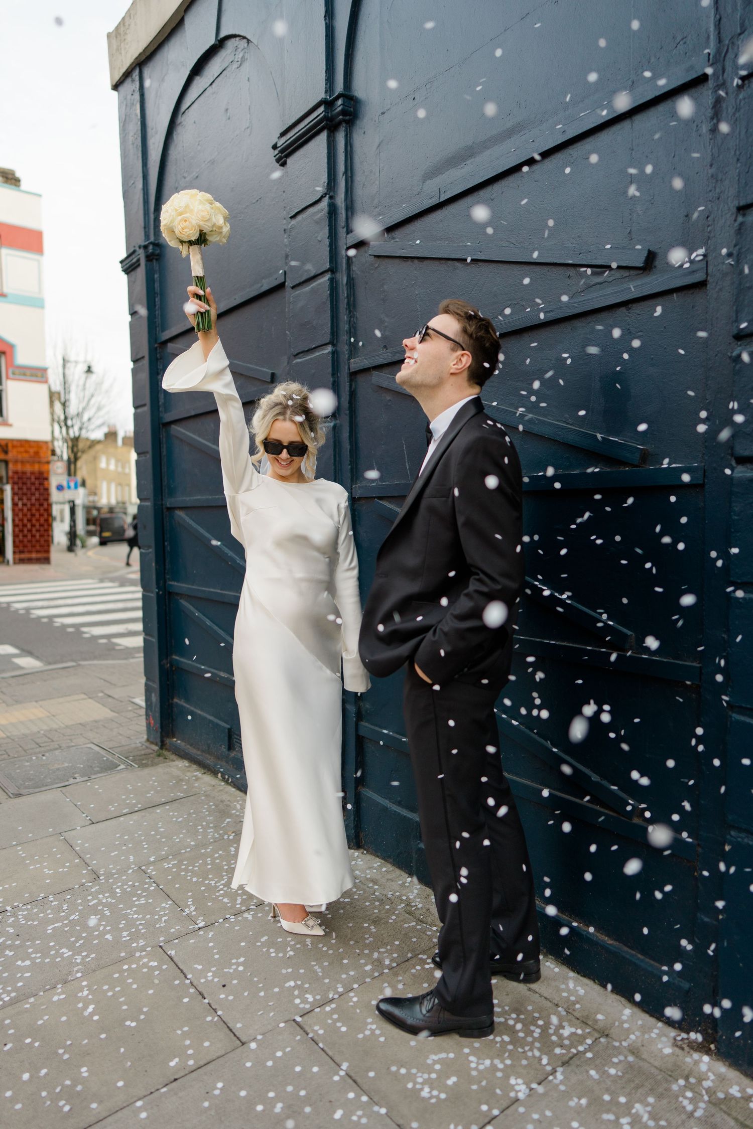 Wedding couple throwing confetti against dark wall during winter snowfall.