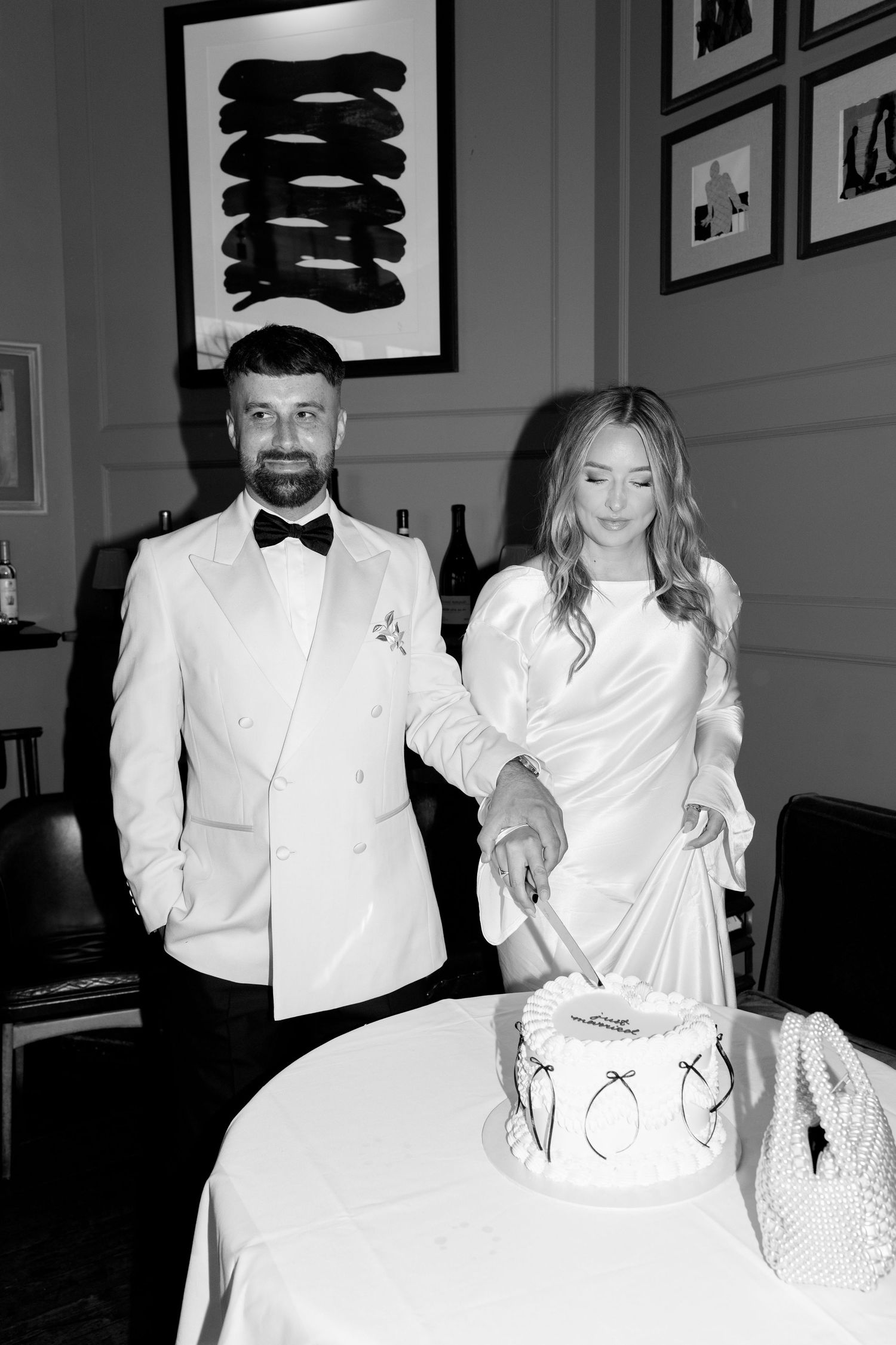 A couple in formal white attire posing together in an elegant indoor setting with framed artwork.