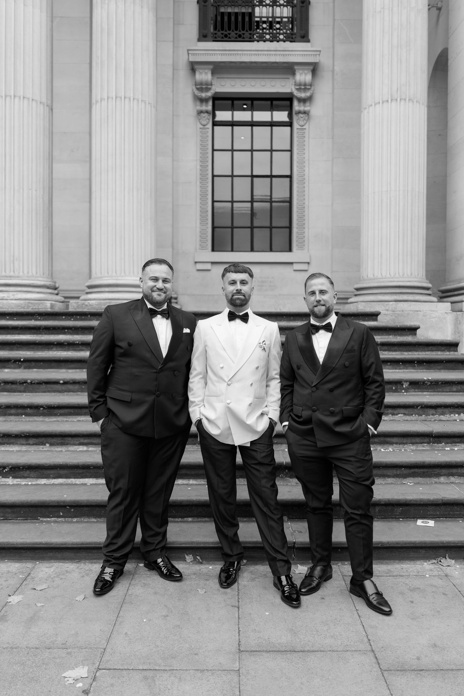 Groomsmen in formal attire sit on steps of classical building.