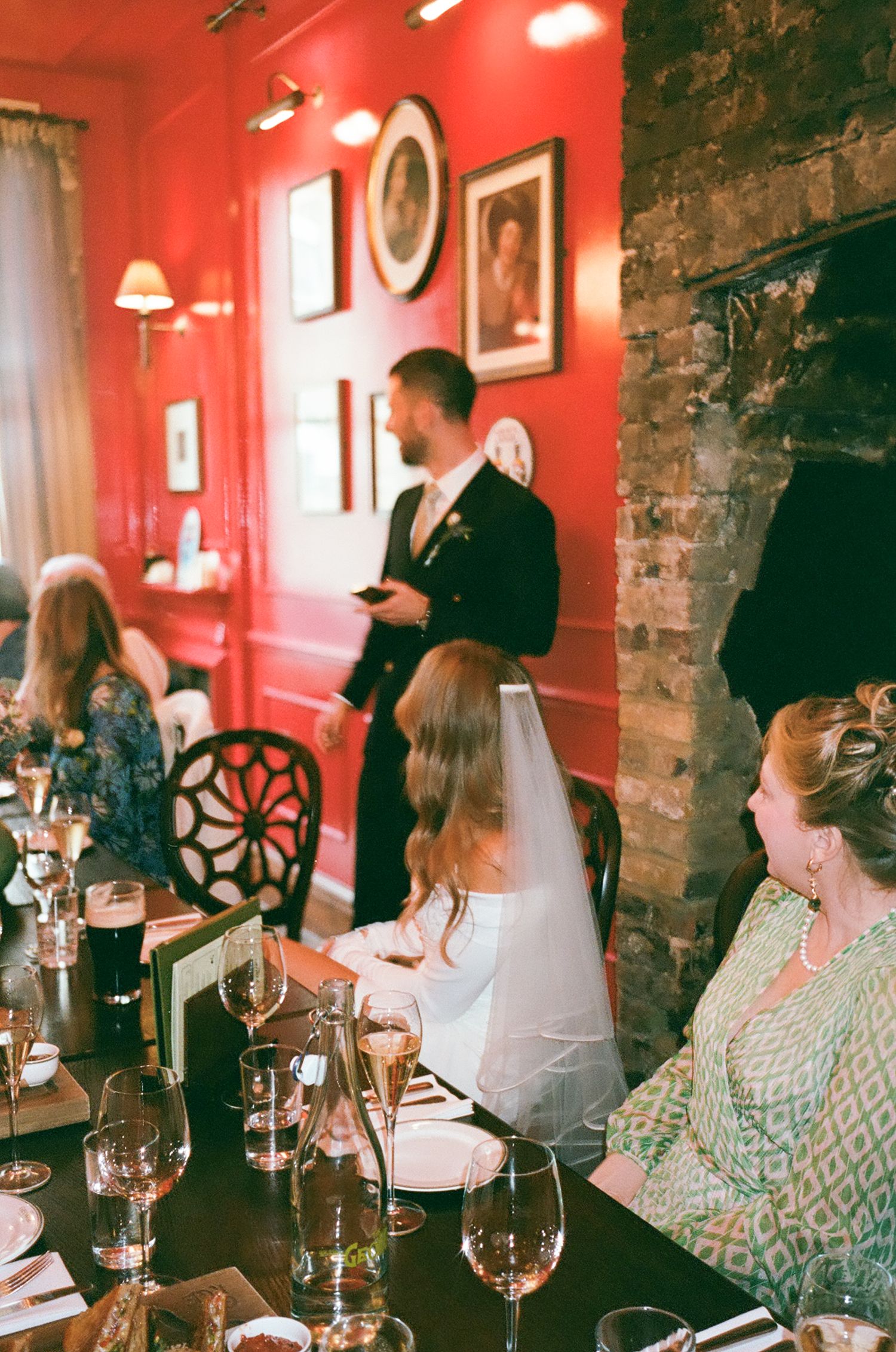 Guests seated at a restaurant table with red walls and exposed brick enjoying wine and conversation in warm lighting.