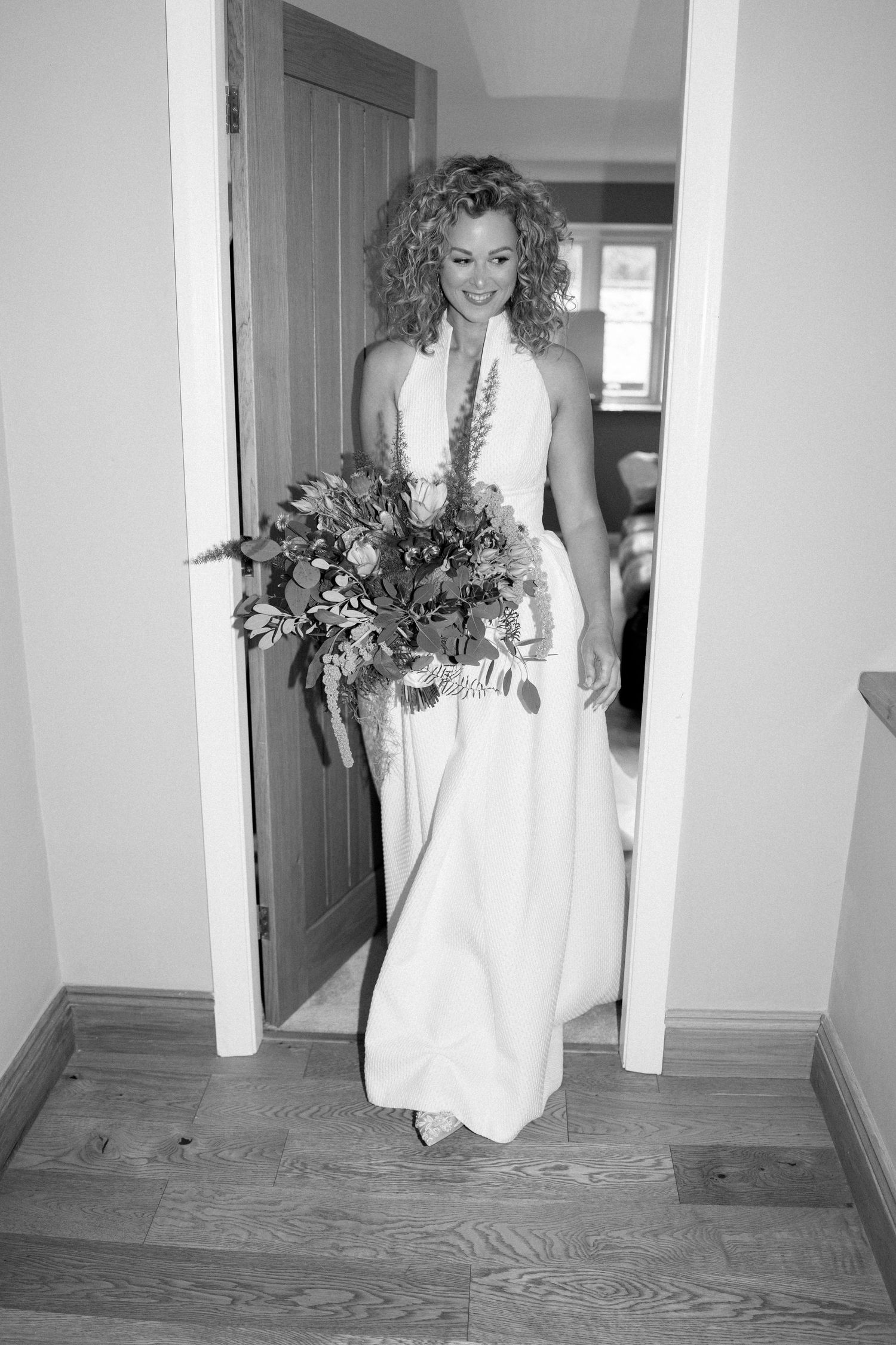 Person in a white wedding dress walking through a hallway carrying a bouquet.
