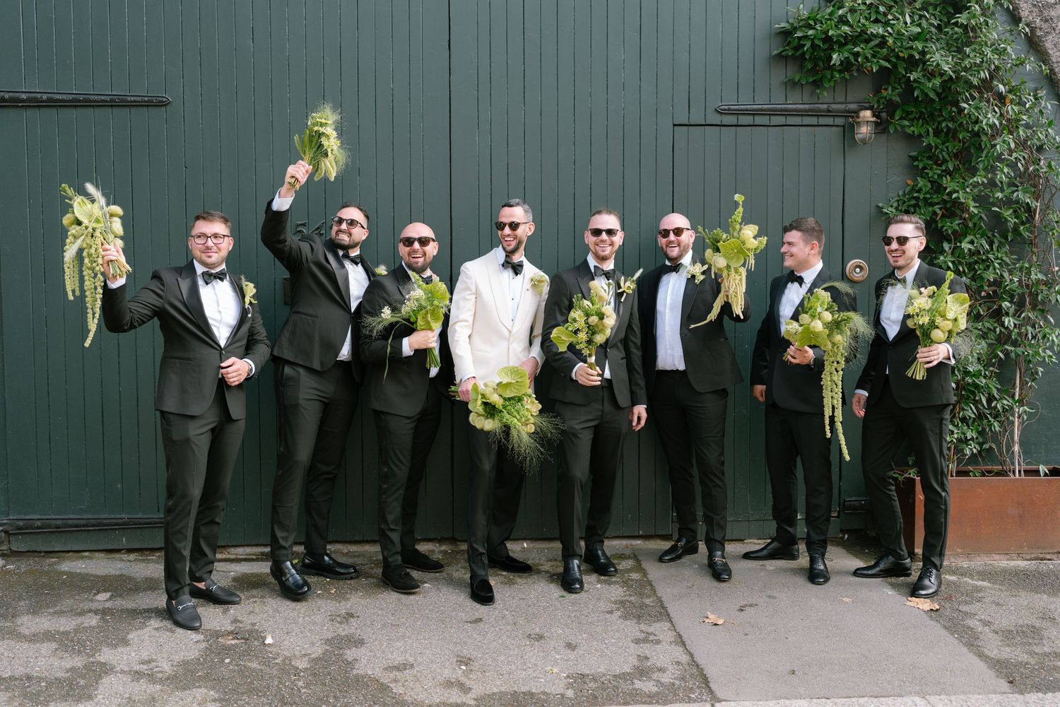 Wedding party in black suits and green bouquets posed against dark green wall outdoors.