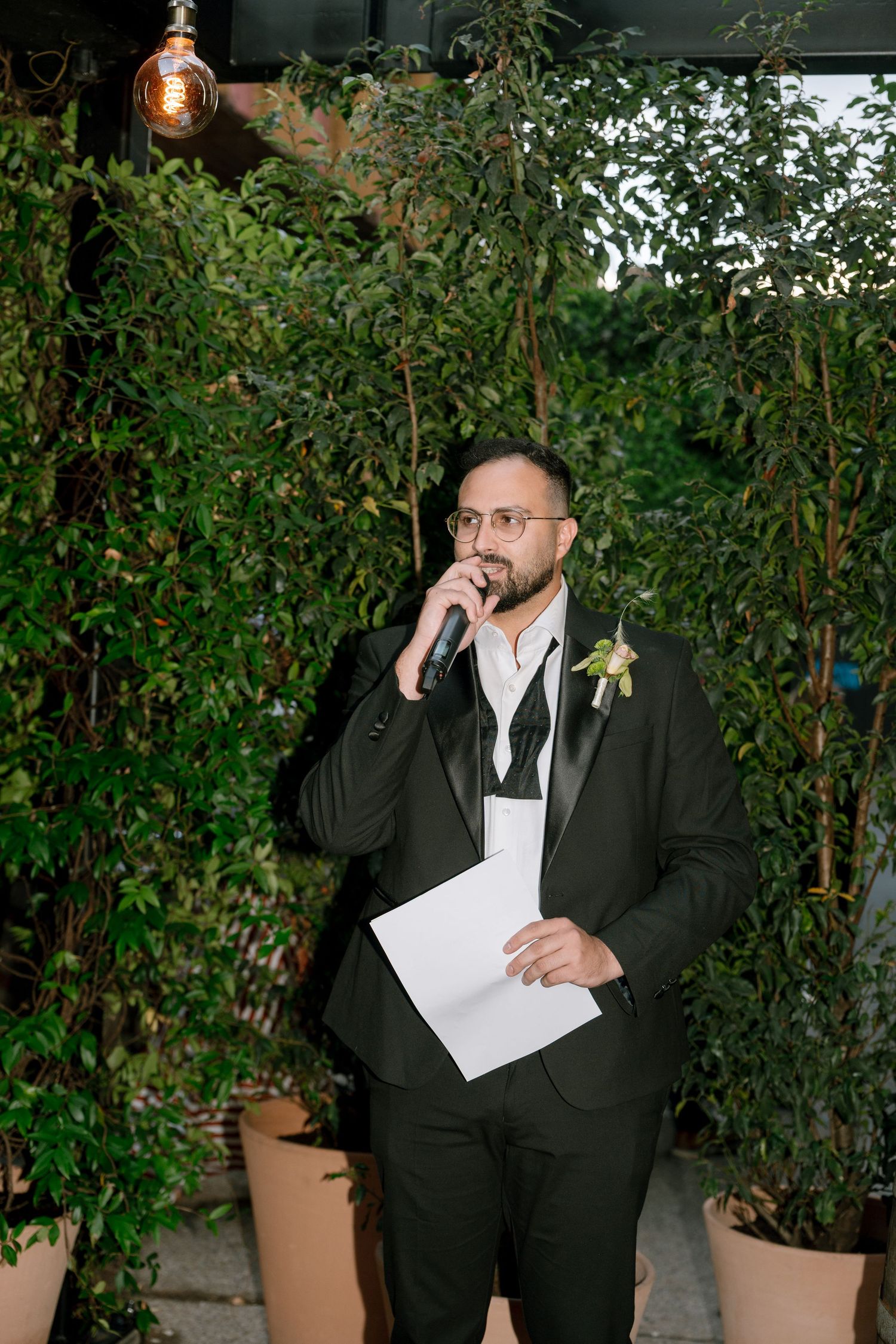 Wedding guest in formal attire giving a speech in an outdoor garden setting.