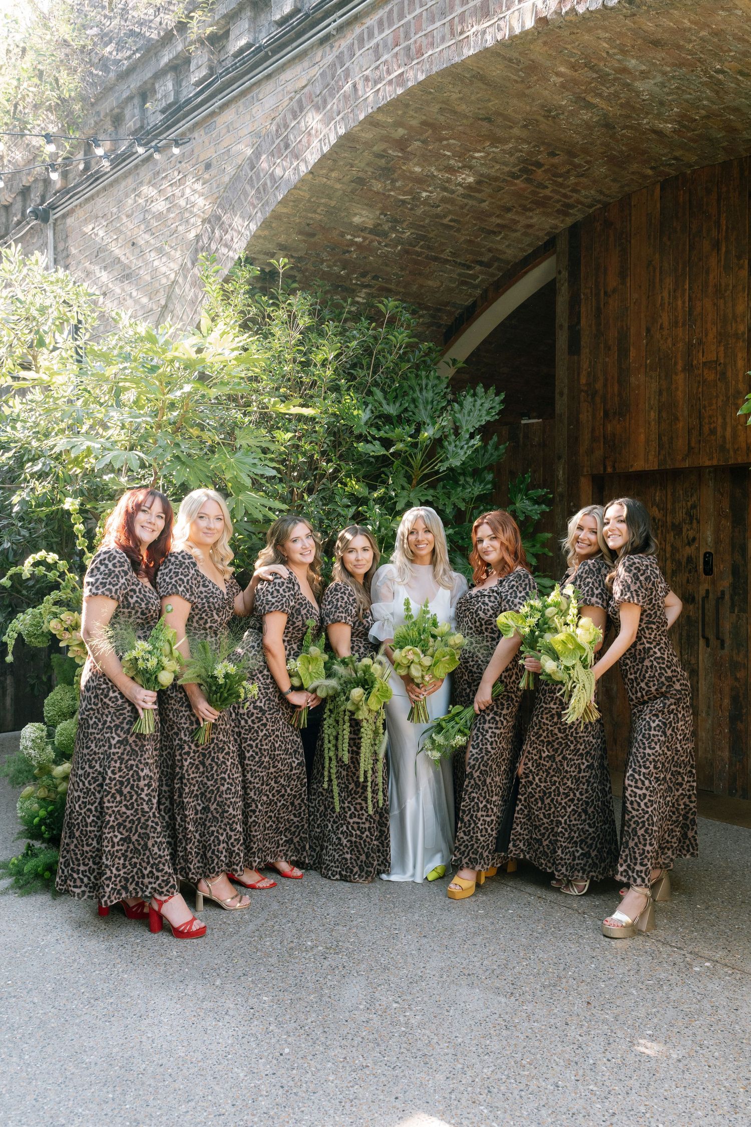 Bridesmaids in matching leopard print dresses pose together under brick archway.