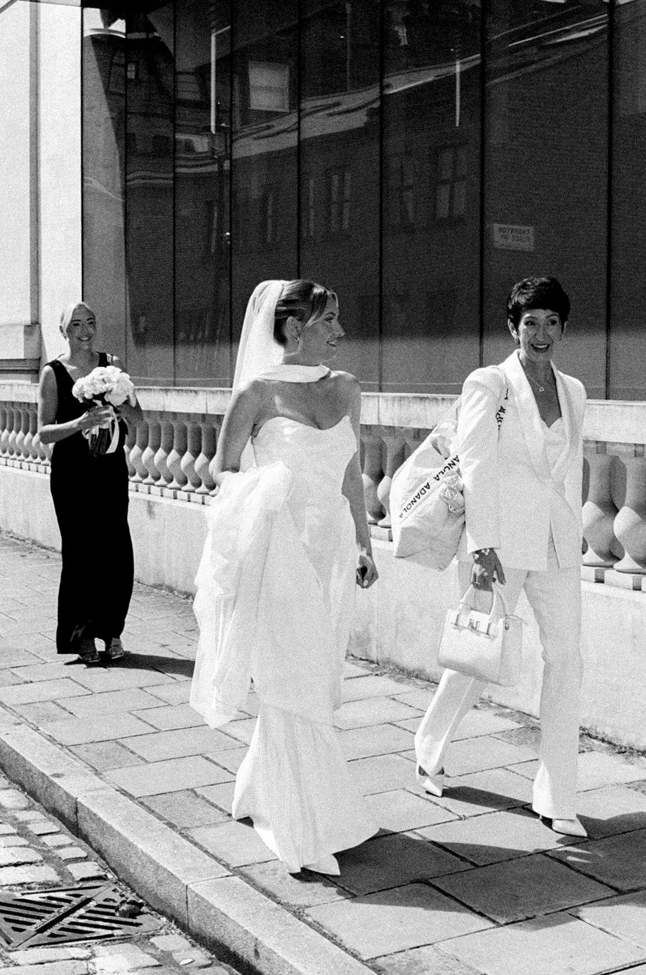Black and white photo of a bride in white gown walking with guests on a cobblestone street on her wedding day.
