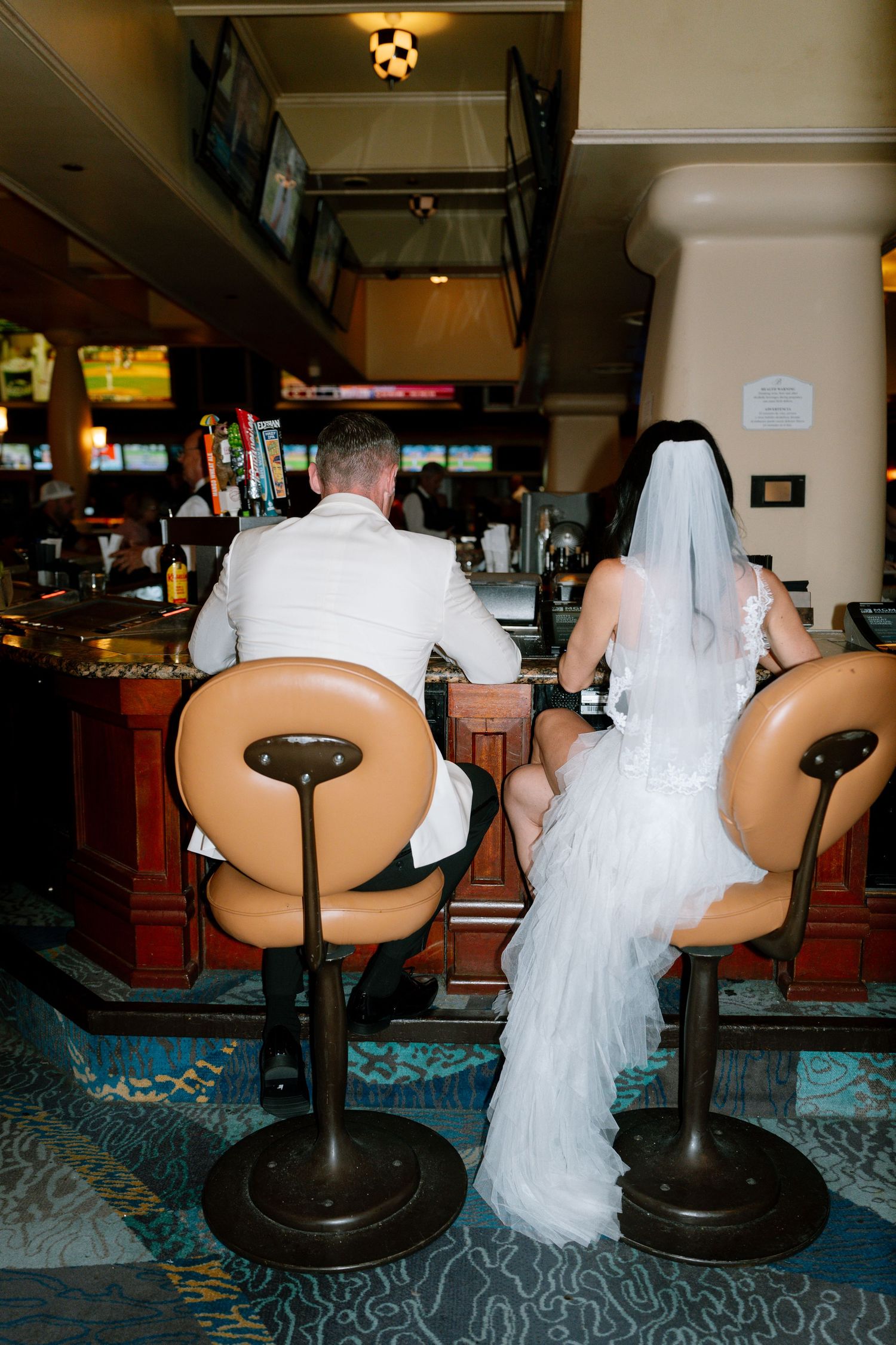 Rear view of couple sitting on retro bowling alley seats with white wedding dress flowing.