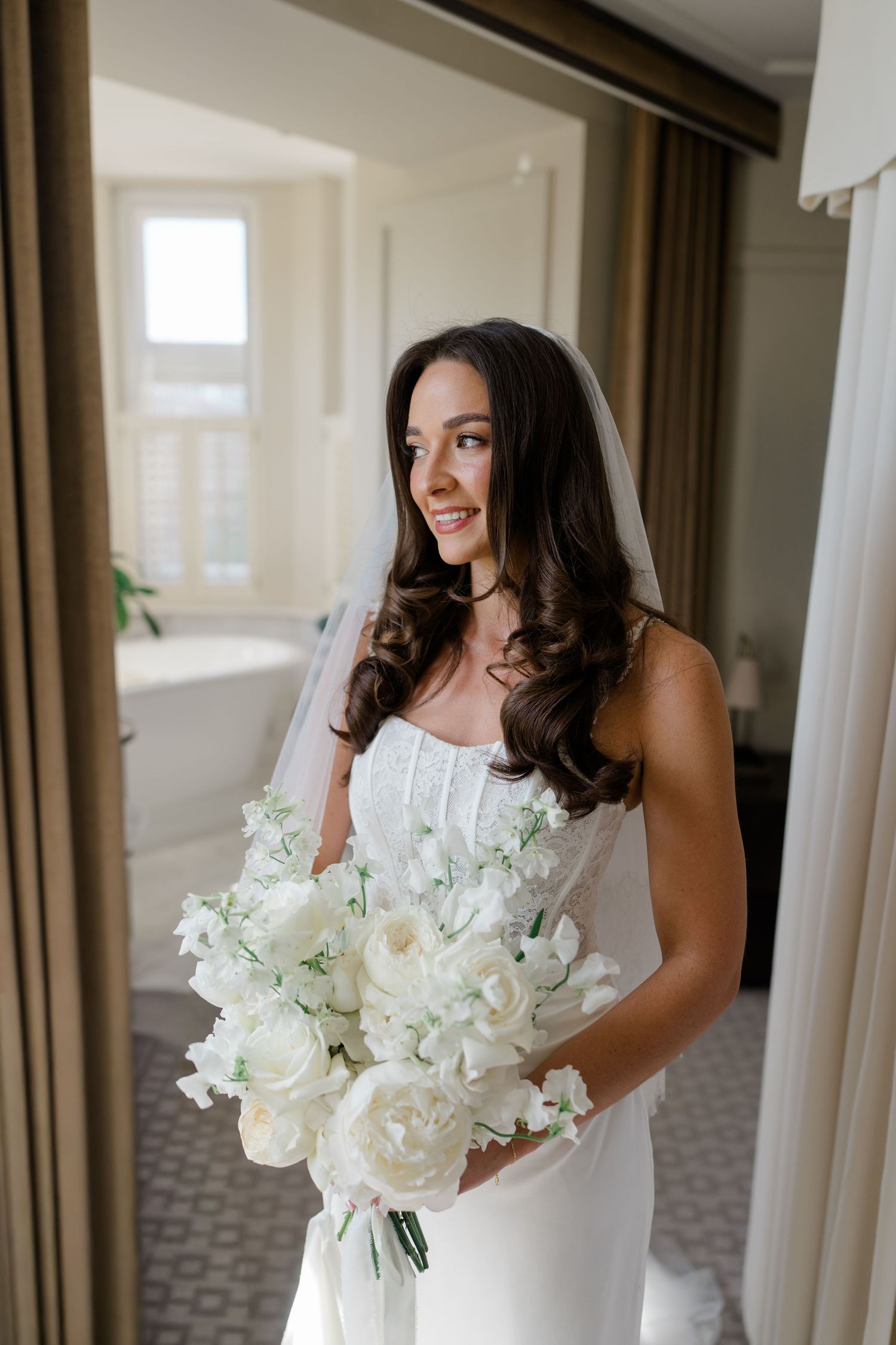 Elegant bride in strapless white gown and veil holds lush white peony and lily bouquet in bright hotel suite.