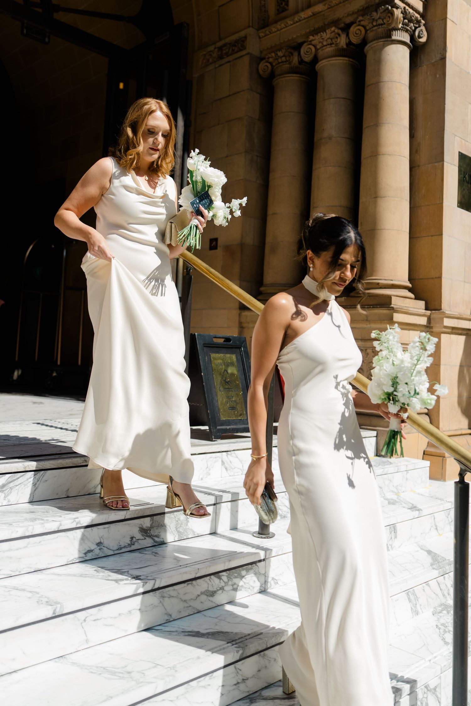 Two bridesmaids in white satin gowns descending marble church steps holding white floral bouquets.