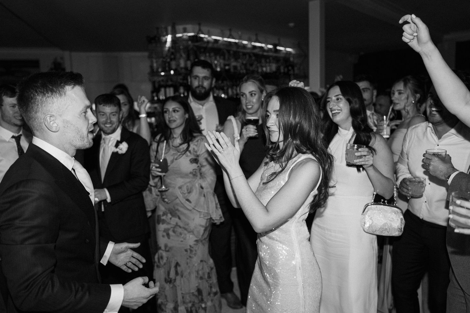 Black and white photo of bride and guests dancing and clapping joyfully at lively wedding reception.