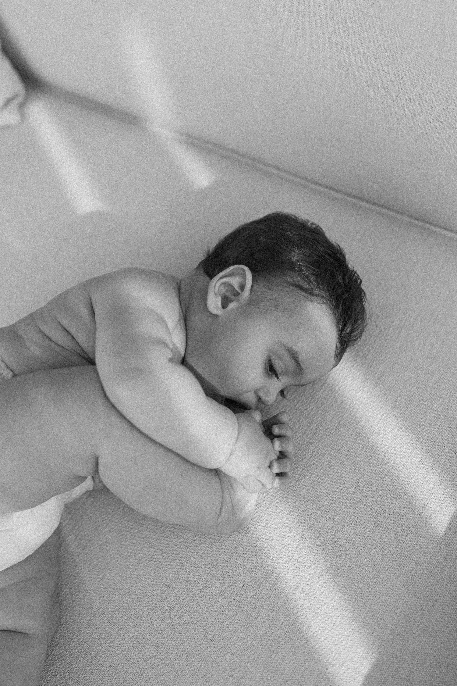 Black and white photo of a sleeping newborn baby lying on a soft textured surface in natural light.