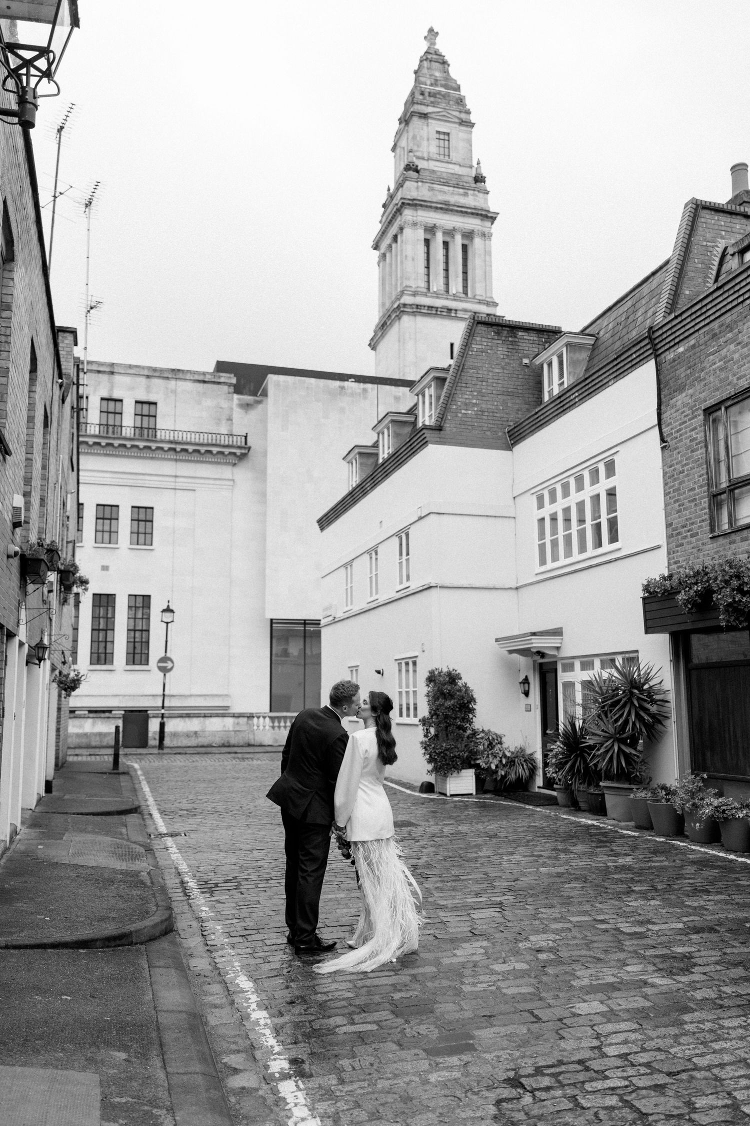 Black and white photo of bride and groom kissing on cobblestone London street.