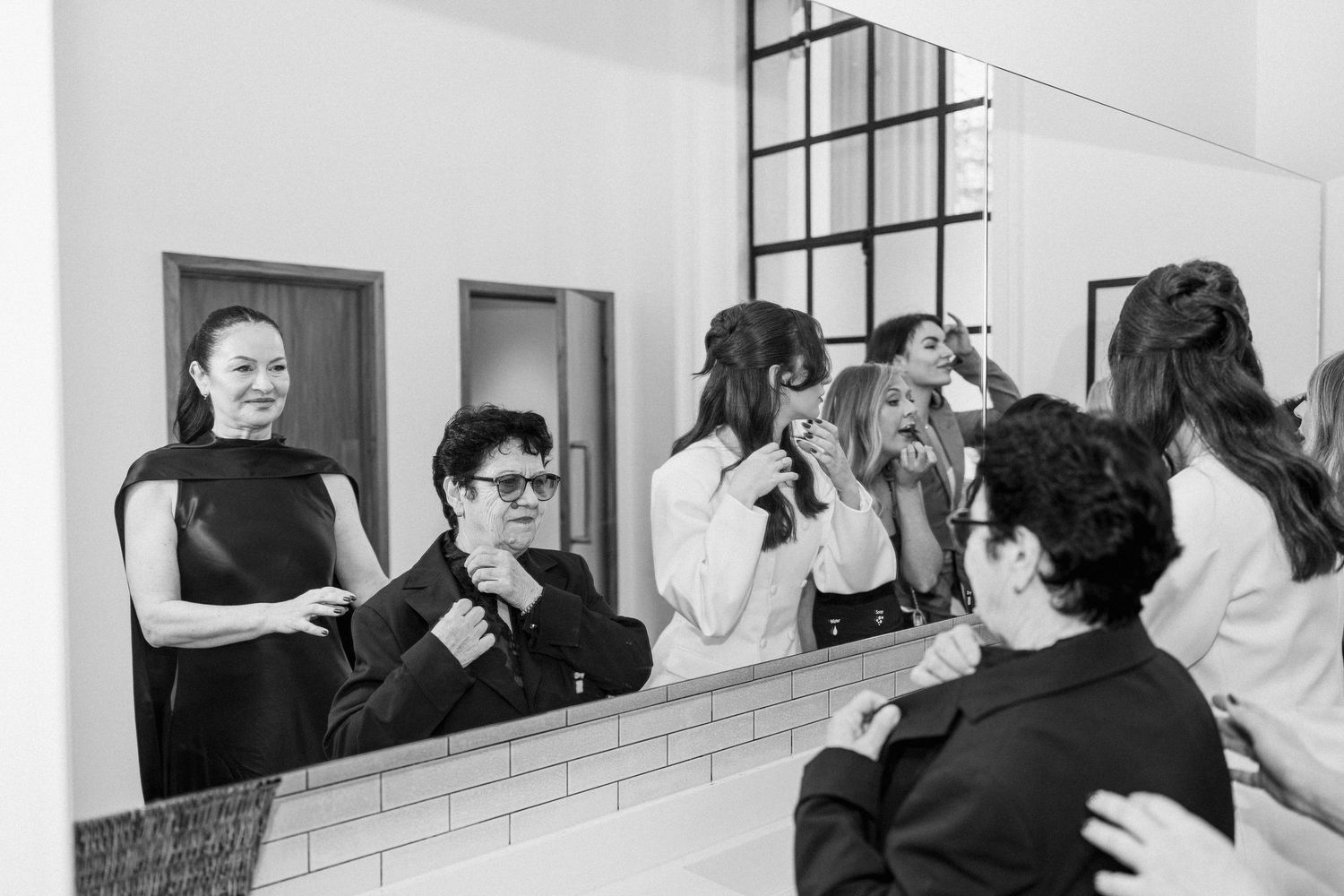 Black and white photo of wedding party laughing and adjusting outfits in front of large bathroom mirror.