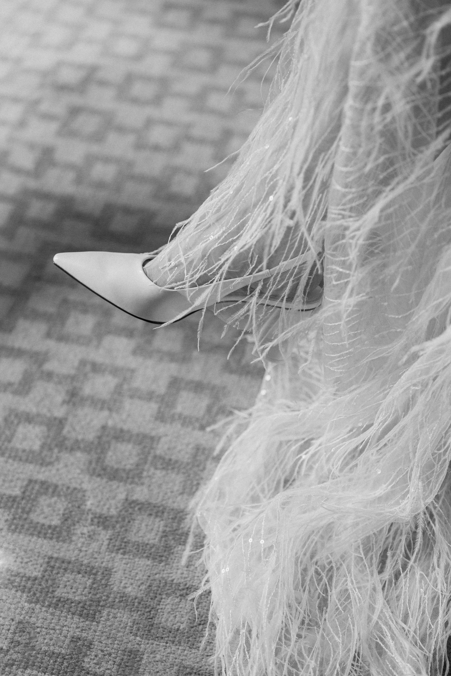 Close-up black and white shot of bridal feathered skirt hem and pointed white stiletto heel.