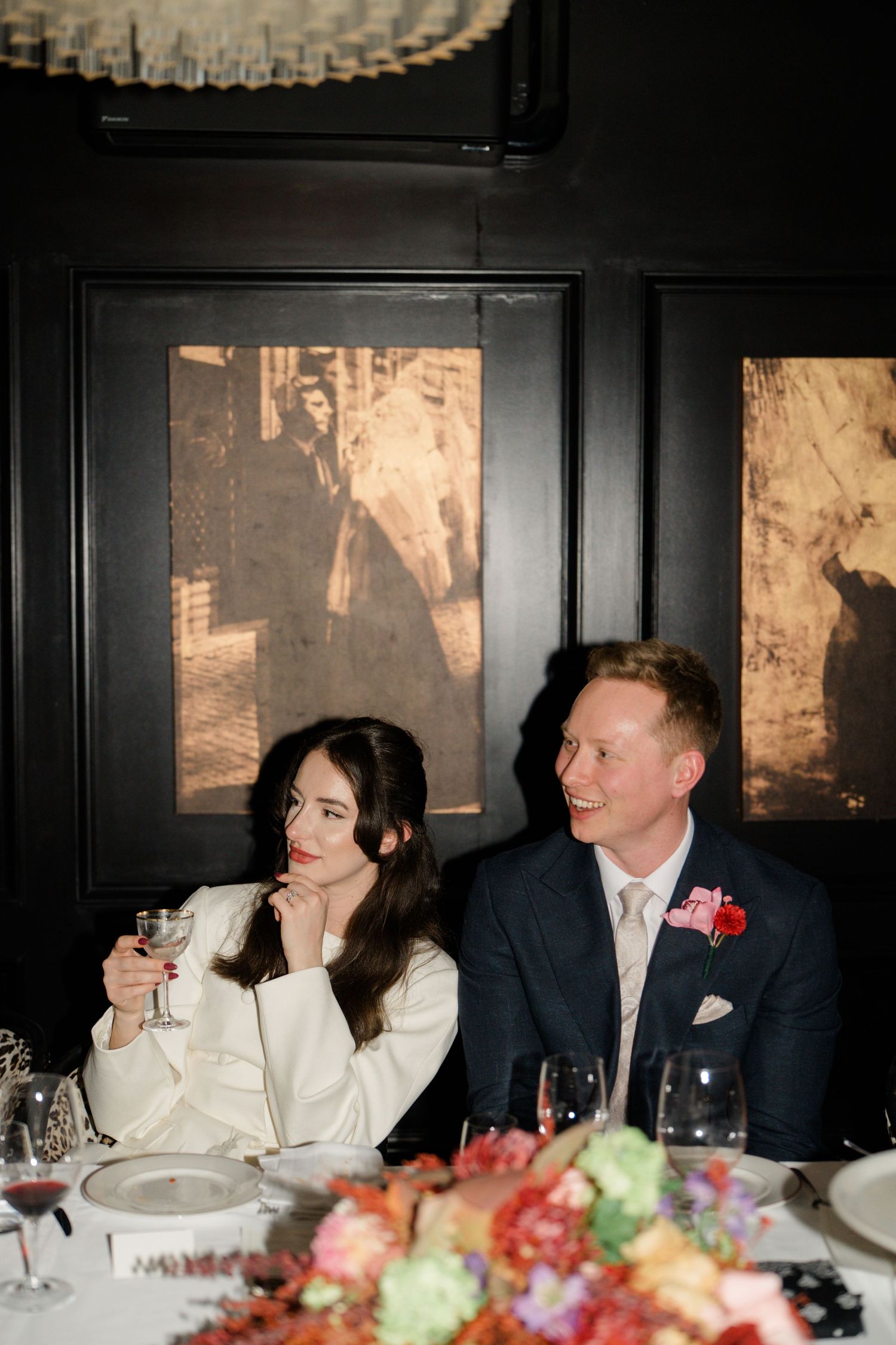Bride and groom sitting at wedding reception table surrounded by colorful floral arrangements.