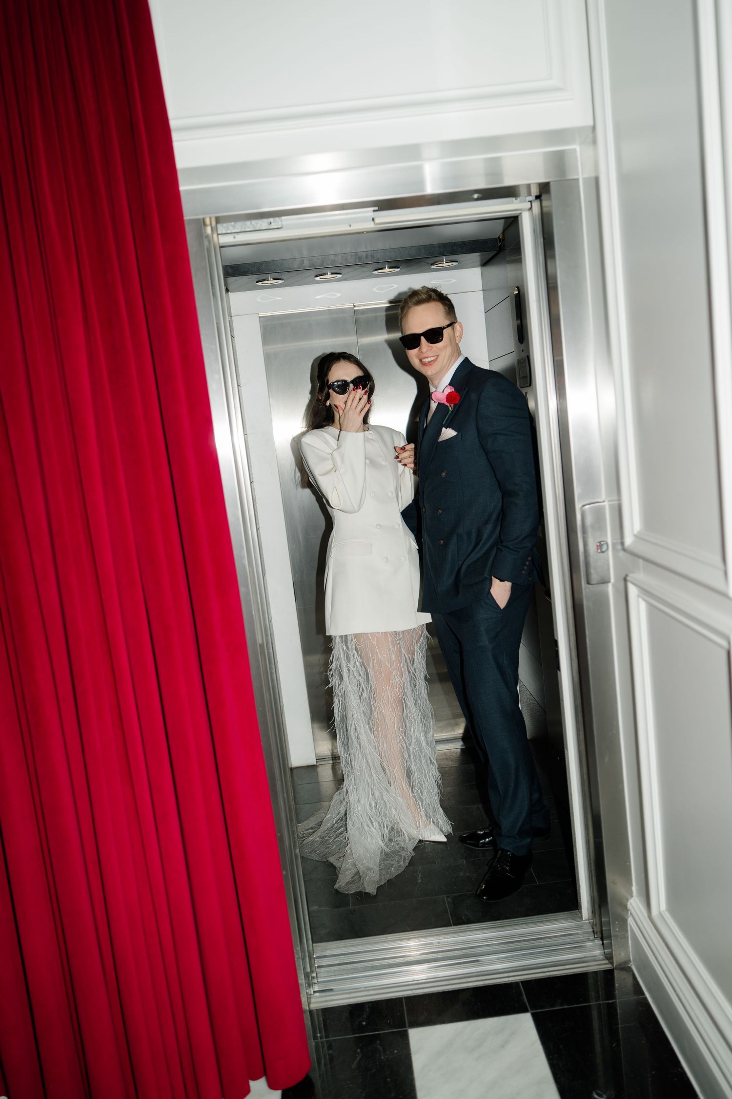 Stylish newlyweds in sunglasses pose in elevator, bride in feather dress, groom with red boutonniere.