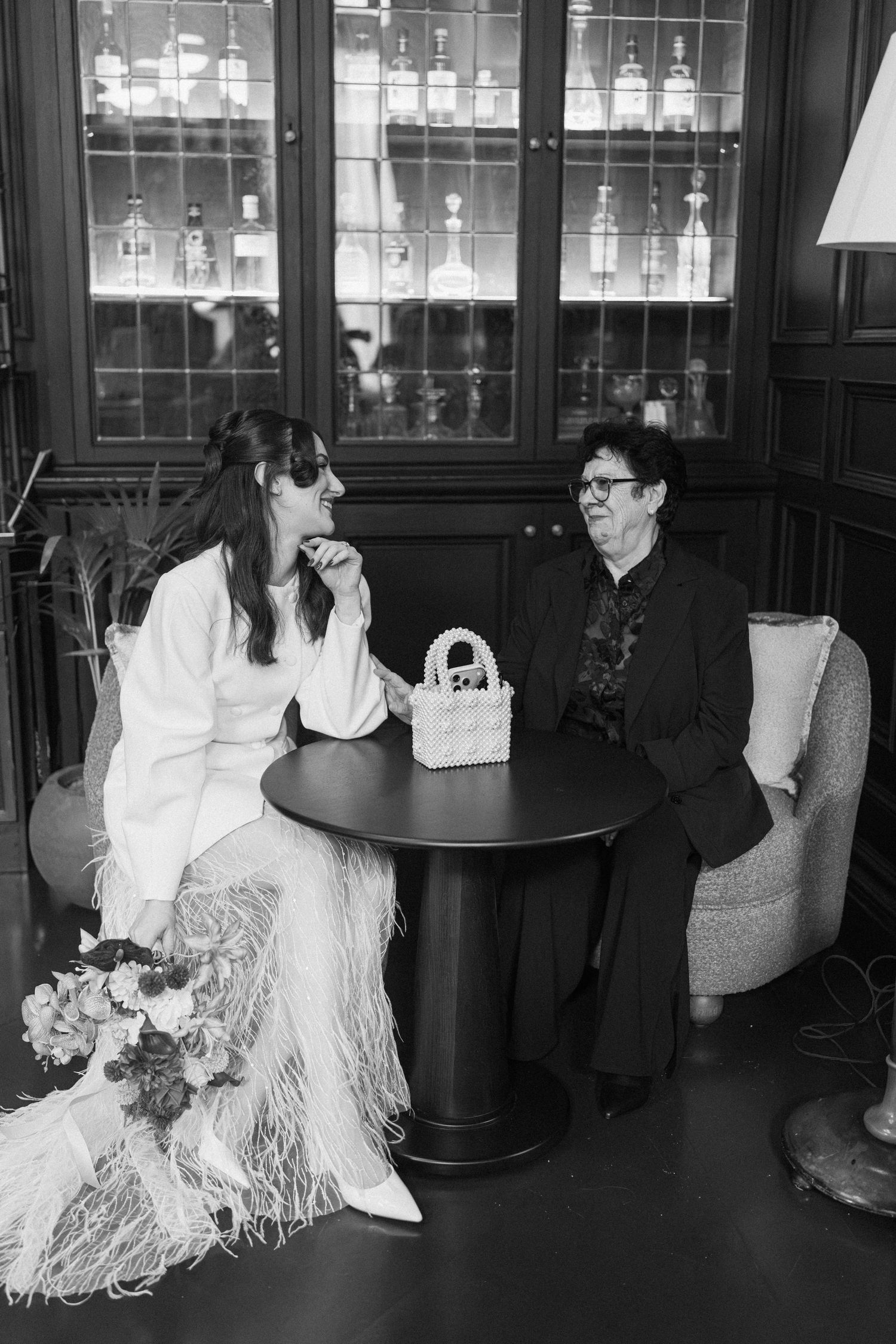 Black and white photo of a bride in feather skirt chatting with an older woman at a round table.