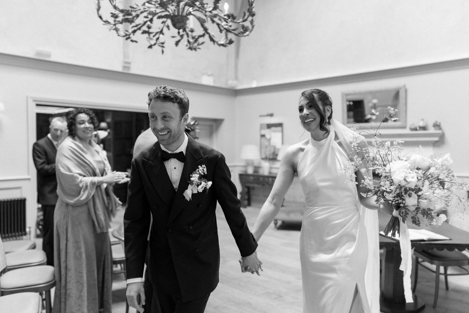 Bride and groom smile holding hands while walking through elegant wedding venue with chandelier overhead.