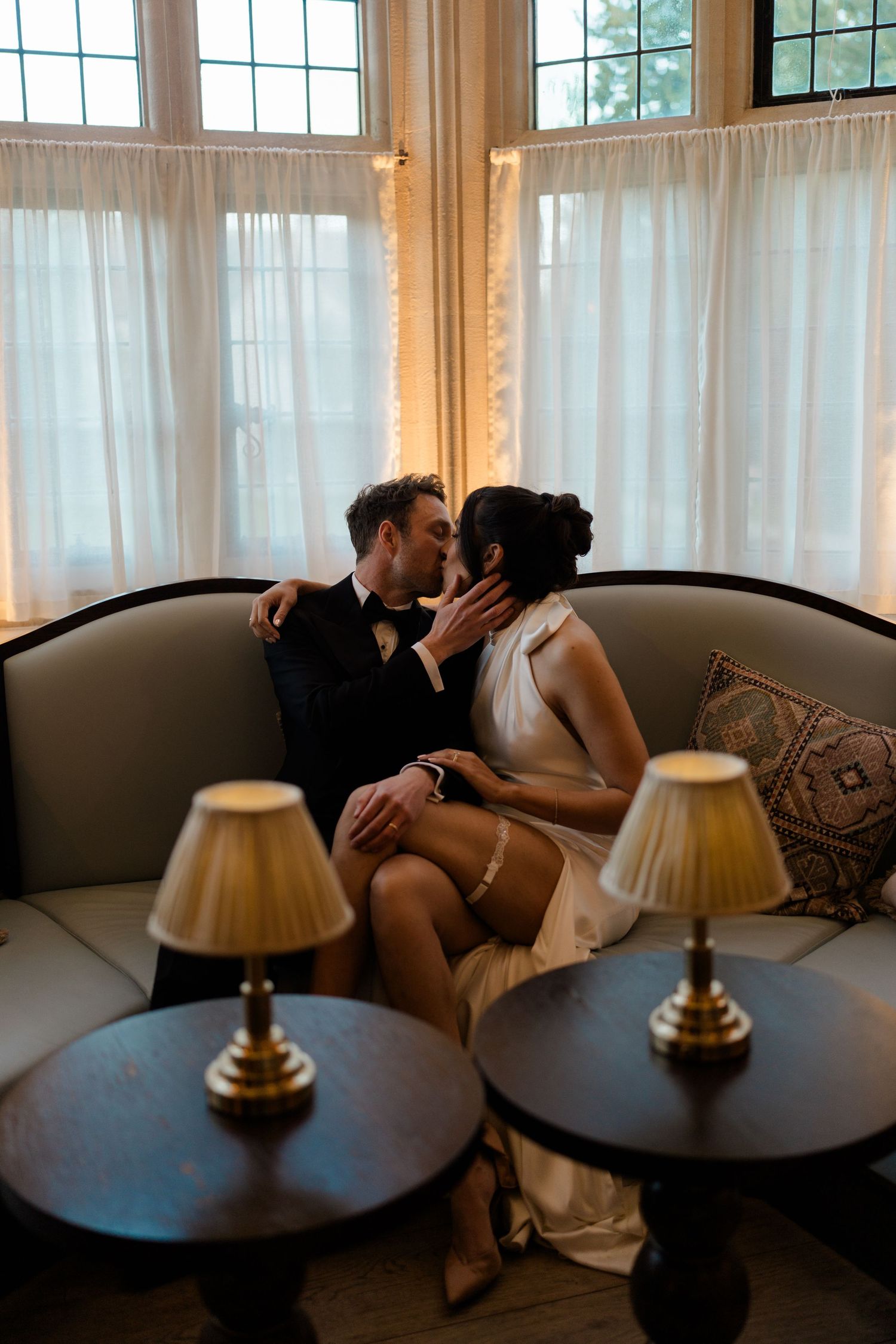 Bride and groom sharing a romantic kiss on a curved sofa by large windows with sheer curtains.