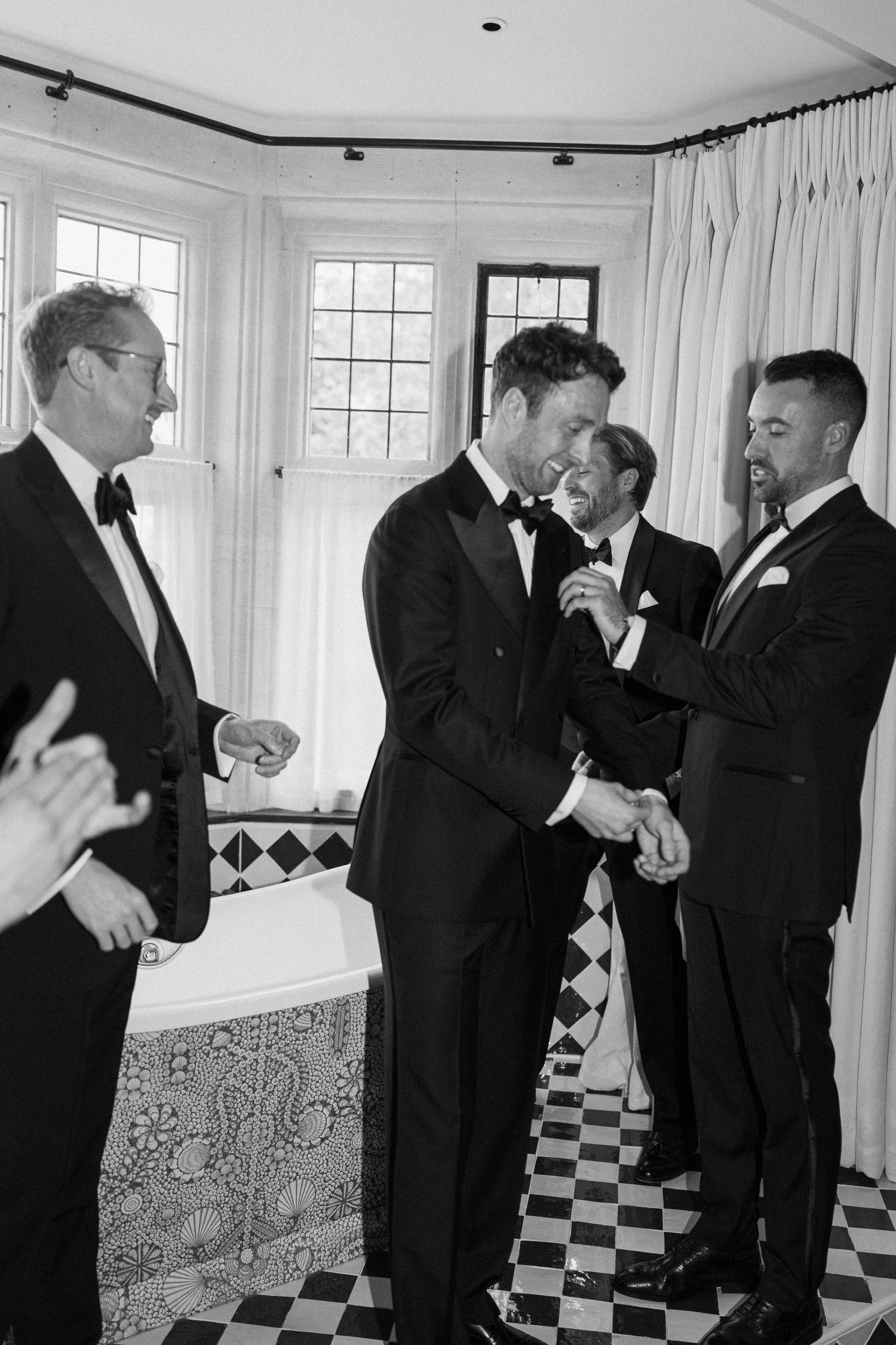 Black and white photo of grooms sharing a joyful moment with groomsmen in tuxedos.
