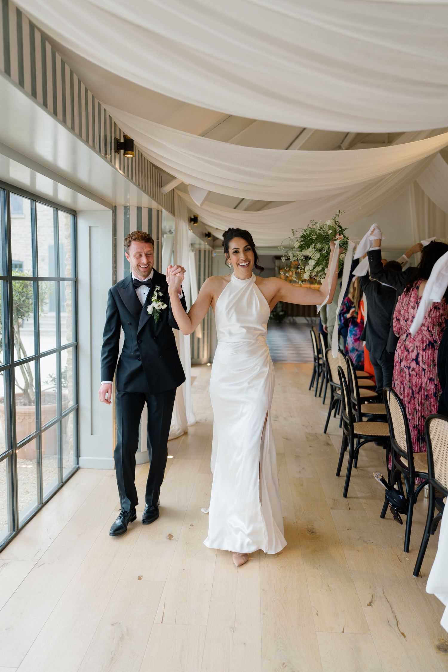 Joyful bride in white halter gown and groom in black tuxedo making grand entrance at elegant wedding reception.