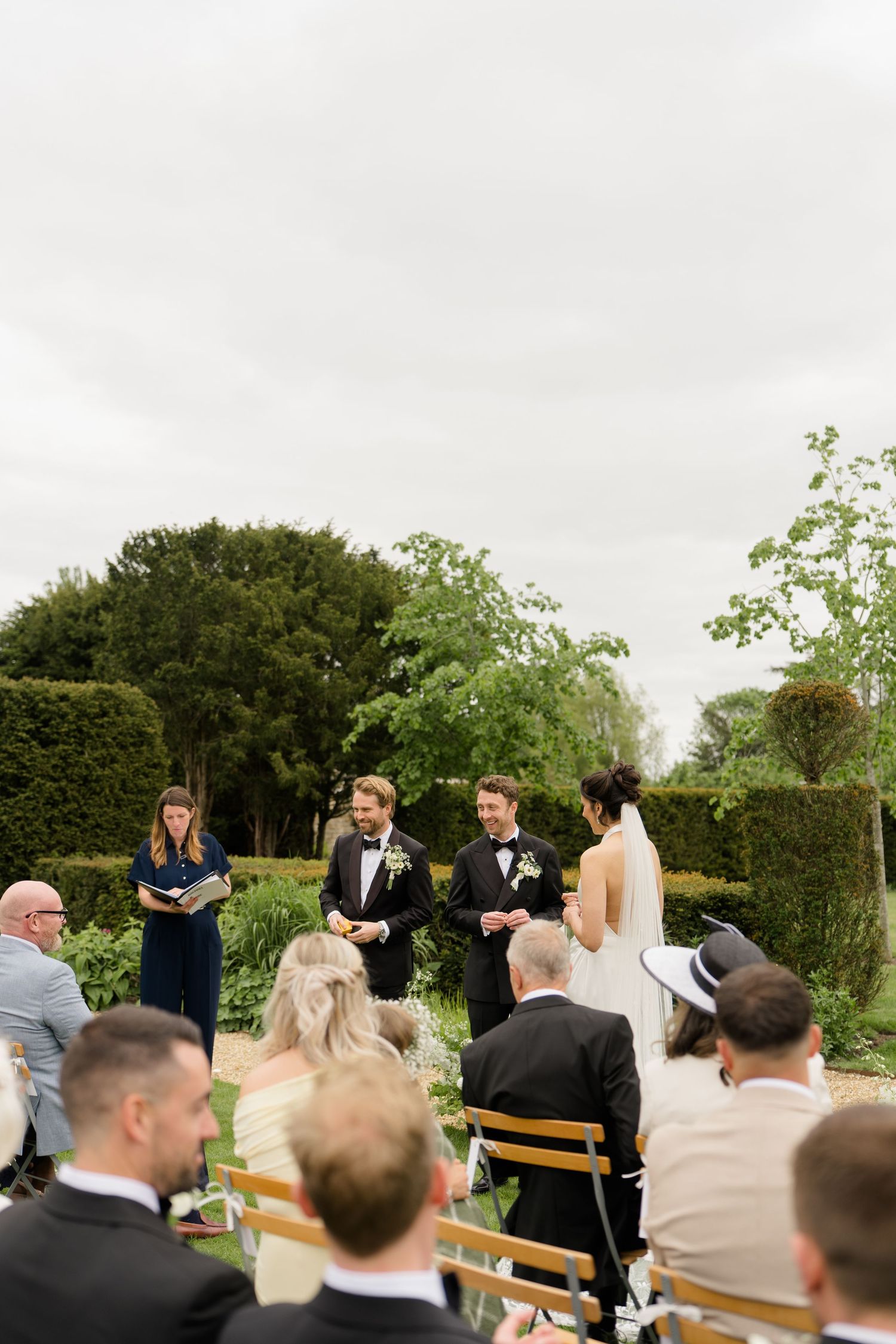 Bride and groom exchange vows in lush garden ceremony with guests seated on wooden chairs outdoors.