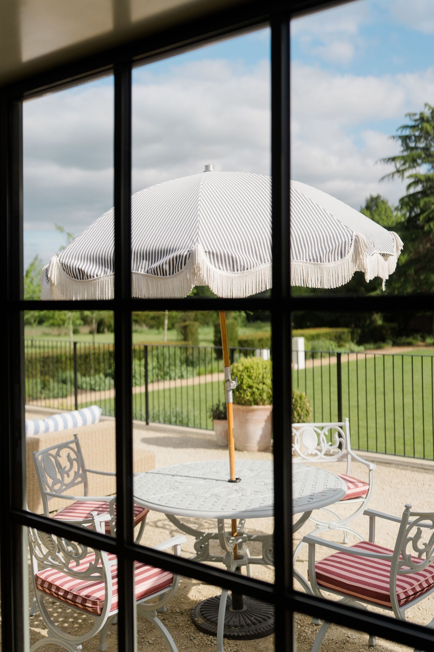 View through window panes of an elegant outdoor patio with striped umbrella and ornate chairs.