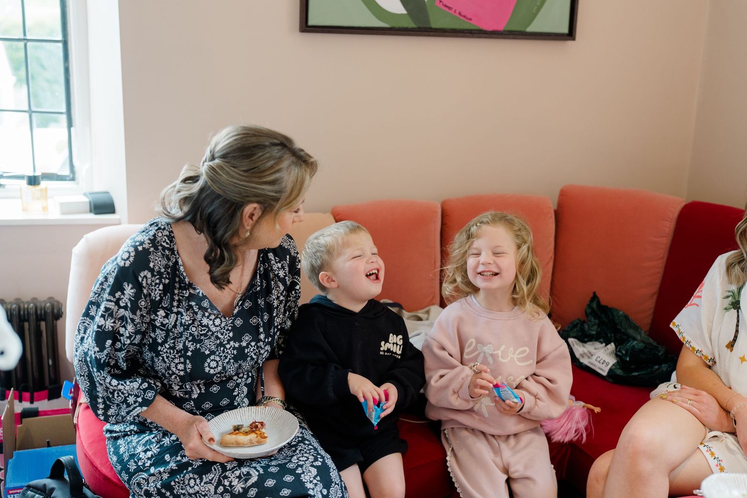 Woman laughing with two young children on a red sofa during wedding morning preparations.