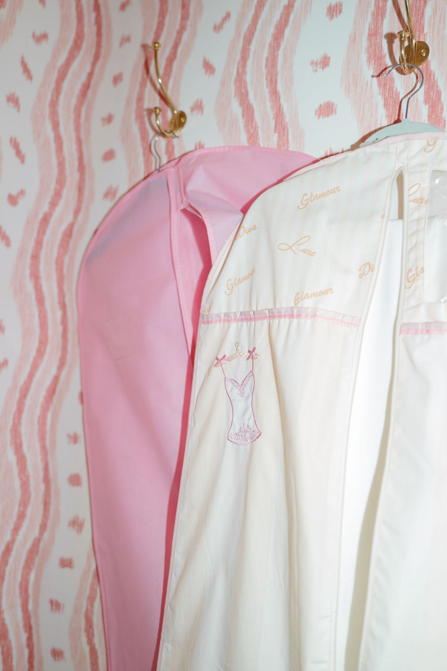 Pink and cream garment bags hanging on gold hooks against pink striped wallpaper in a bridal dressing room.