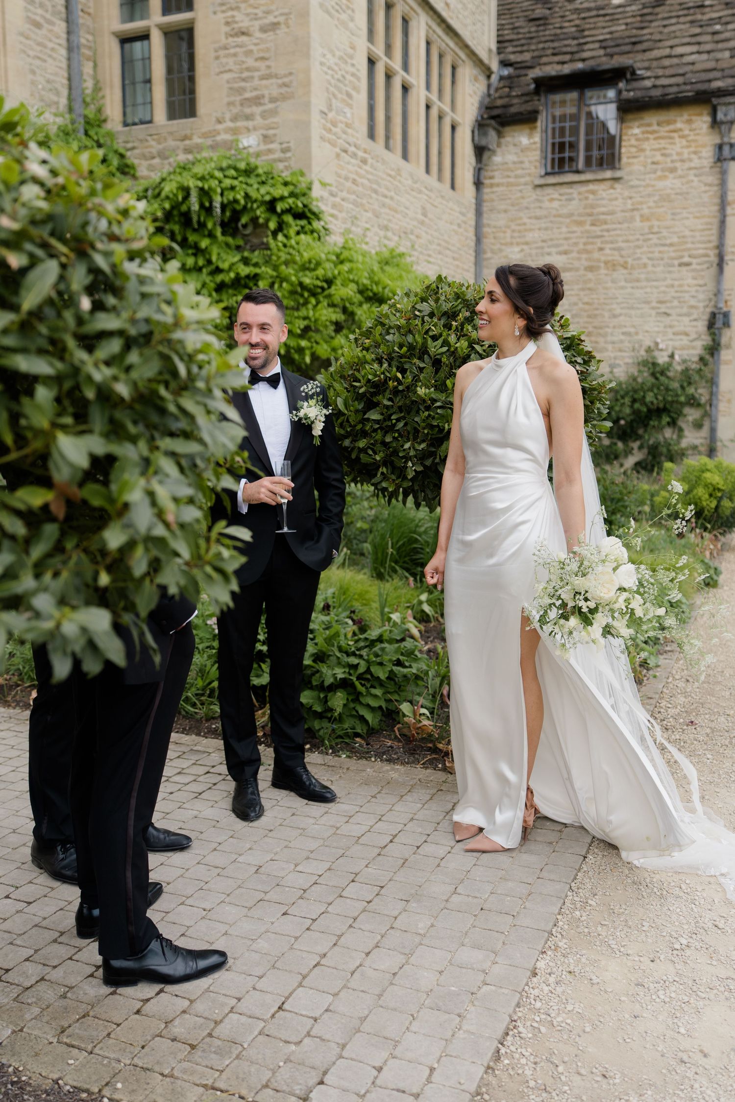 Smiling groom in black tuxedo stands with bride in halter wedding gown outside a stone manor.