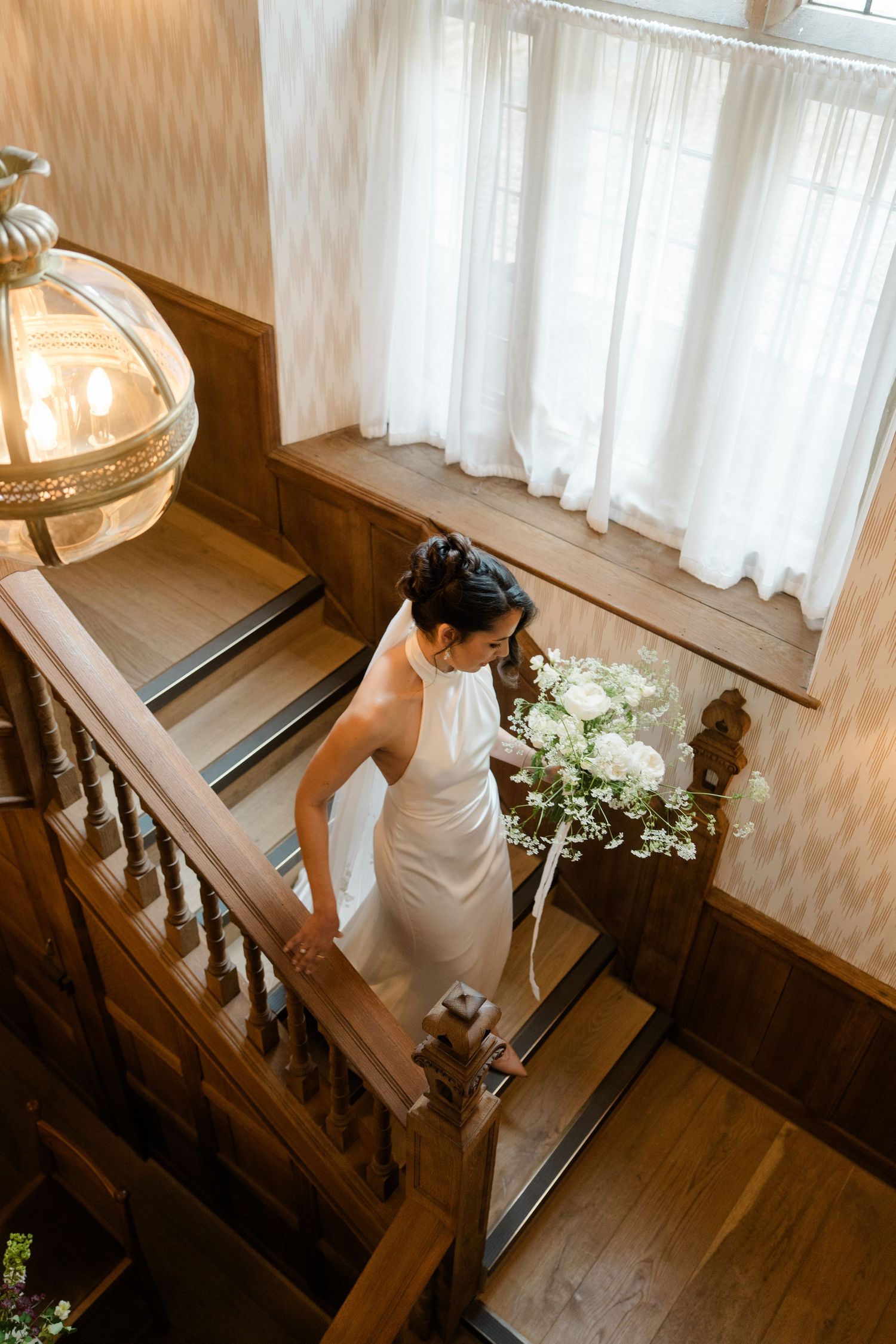Aerial view of bride in sleek white gown descending wooden staircase with white bouquet.