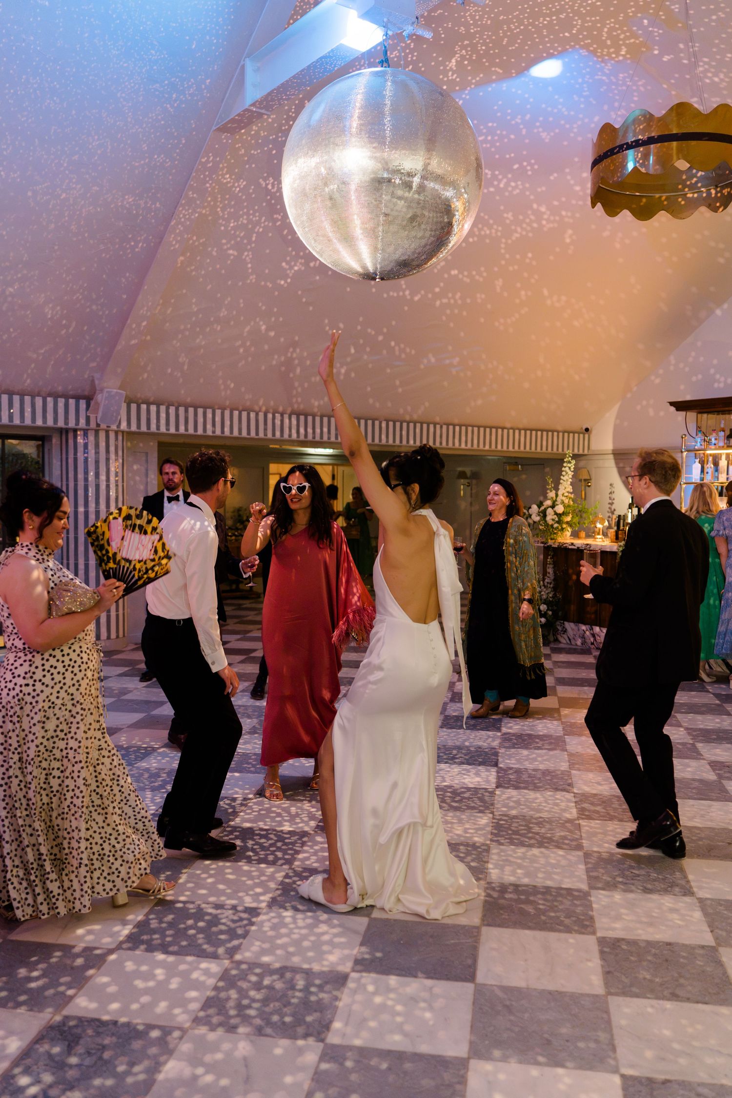Bride in white gown dancing under giant disco ball with guests on checkered dance floor.